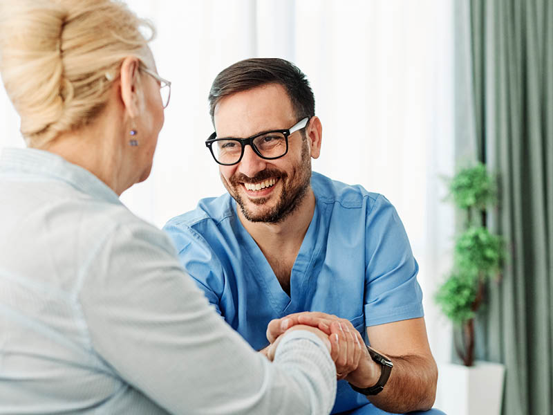 Positive Interaktion zwischen Pflegekraft und Patientin Eine Pflegekraft, erkennbar an den blauen Berufskleidung, hält lachend die Hand einer Frau, deren Rücken zur Kamera zeigt. Die Szene spielt sich in einem hellen Raum ab.