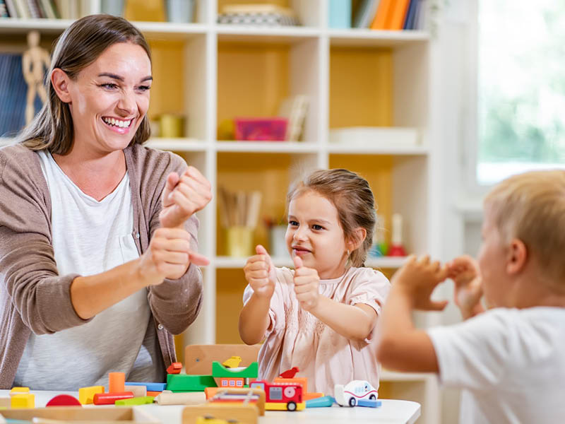 Eine Erwachsene und zwei Kinder spielen mit bunten Bauklötzen an einem Tisch vor einem Bücherregal. Die Erwachsene hält ein Spielzeugstück und gestikuliert, als würde sie den Kindern etwas erklären oder mit ihnen spielen. Die Szene spielt sich wahrscheinlich im Umfeld einer Kindertagesstätte ab und betont Lern- oder Spielaktivitäten.