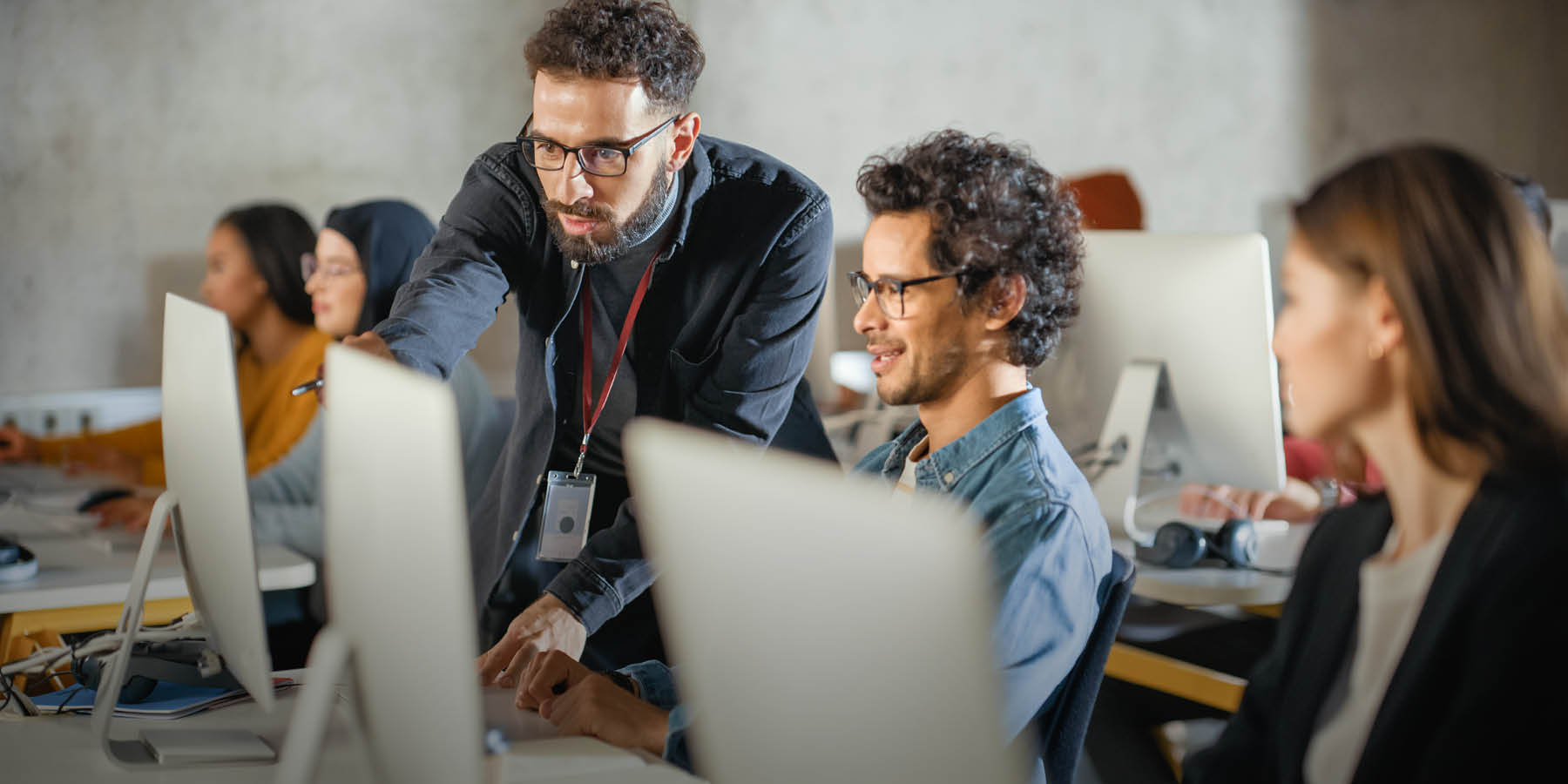 Ein Mann mit Bart und Brille zeigt einem anderen Mann, der ebenfalls eine Brille trägt, etwas auf einem Computerbildschirm. Sie befinden sich in einem modernen Schulungsraum mit weiteren Personen, die an Computern arbeiten.