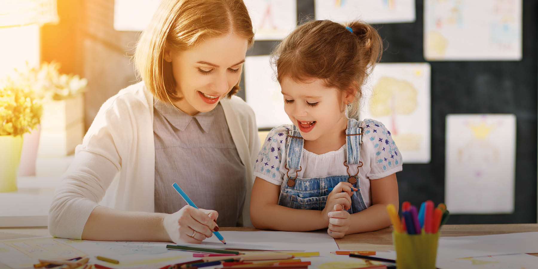 Eine lächelnde junge Frau sitzt an einem Tisch und zeichnet zusammen mit einem kleinen Mädchen, das ebenfalls lächelt. Im Vordergrund sind bunte Stifte und Papier zu sehen, im Hintergrund hängen Kinderzeichnungen an einer Tafel.