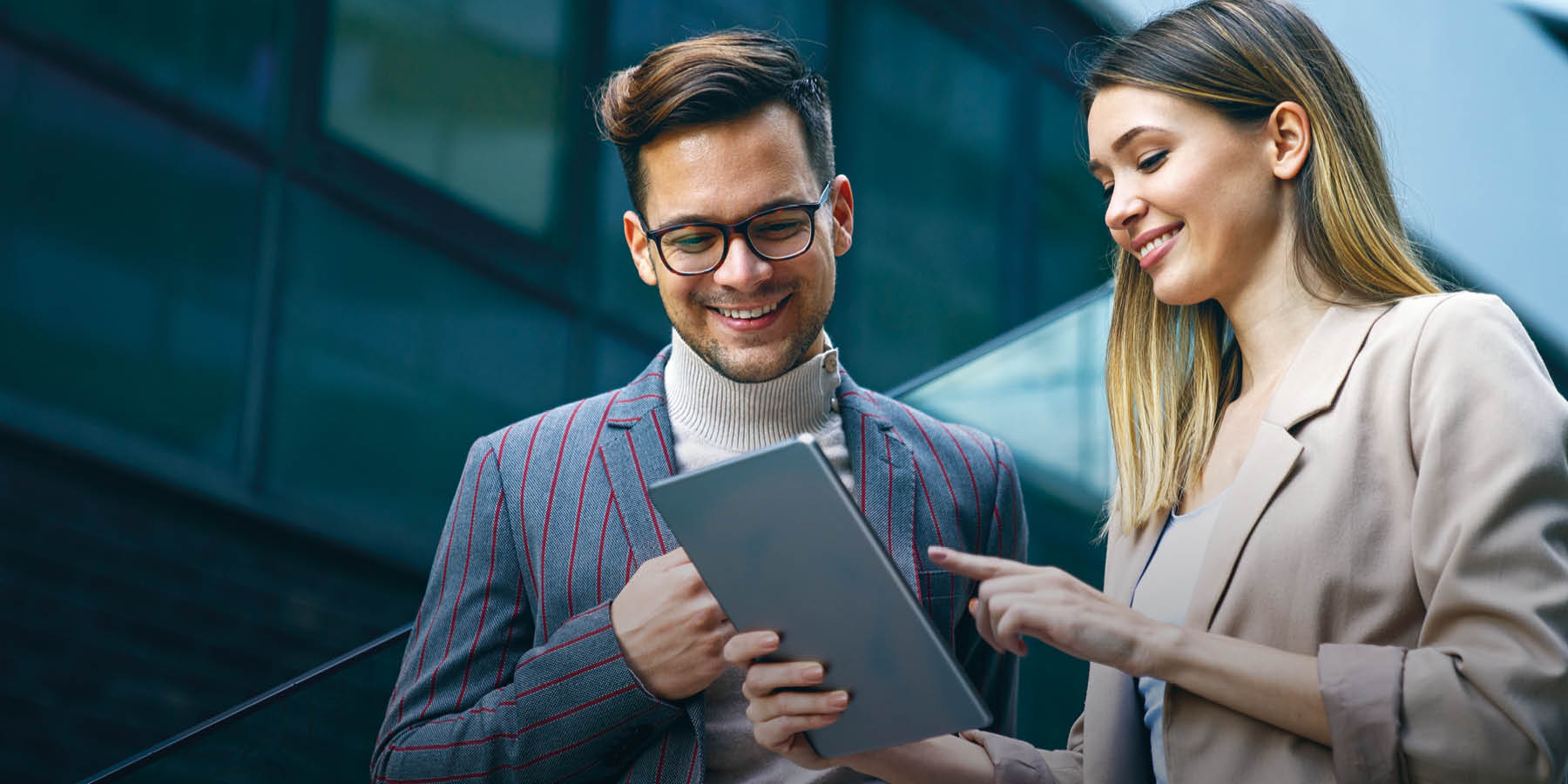 Ein junger Mann und eine junge Frau in geschäftlicher Kleidung stehen im Freien vor einem modernen Gebäude und schauen auf ein Tablet, welches die Frau hält und darauf zeigt. 