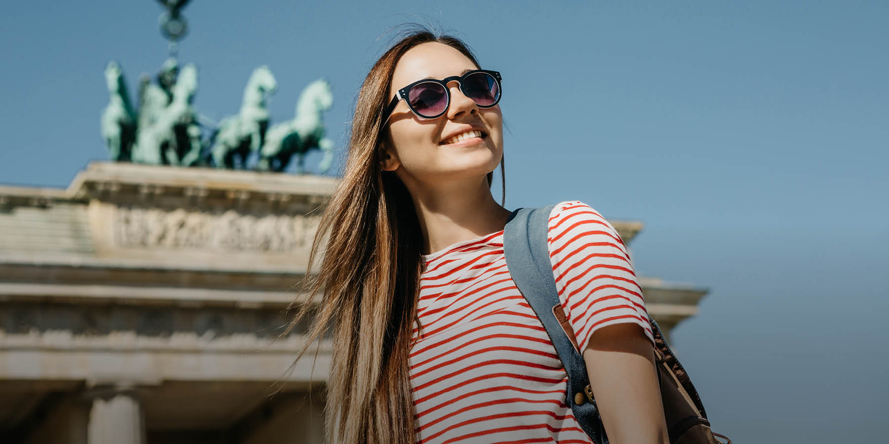 Junge, lächelnde Frau im T-Shirt mit Sonnenbrille und Rucksack steht vor dem Brandenburger Tor.