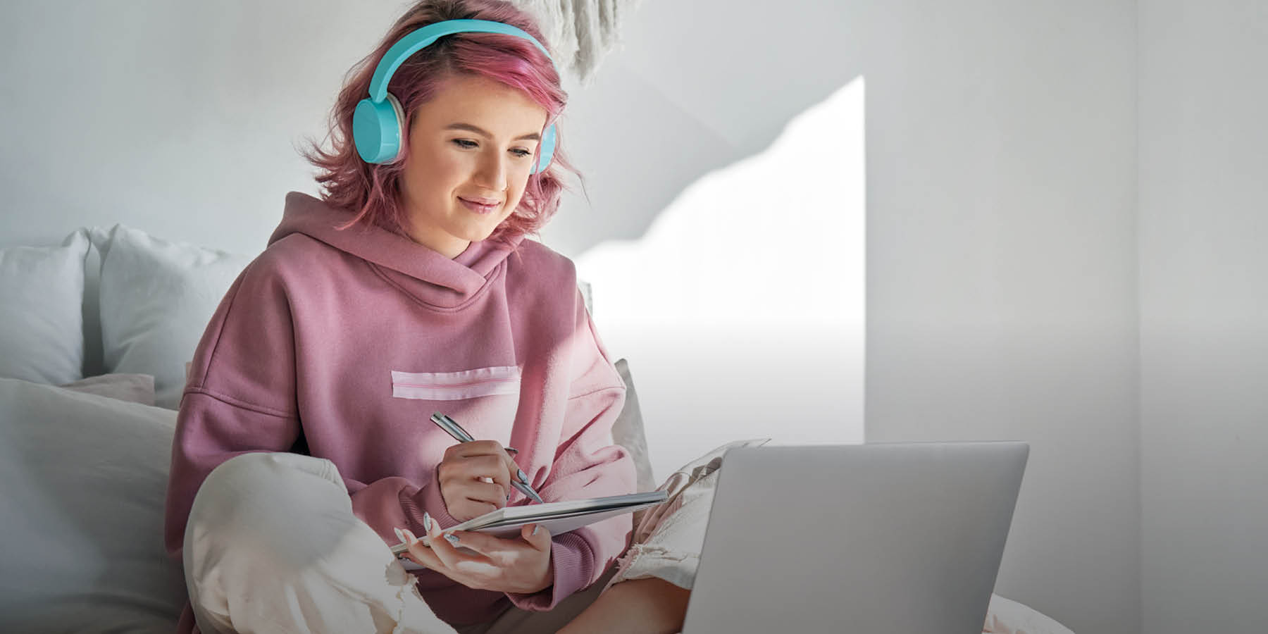 Eine junge Frau mit rosa Haaren, rosa Kapuzenpullover und türkisfarbenen Kopfhörern sitzt im Schneidersitz auf einem Bett vor einem aufgeklappten Laptop und hält einen Notizblock und einen Stift in den Händen.