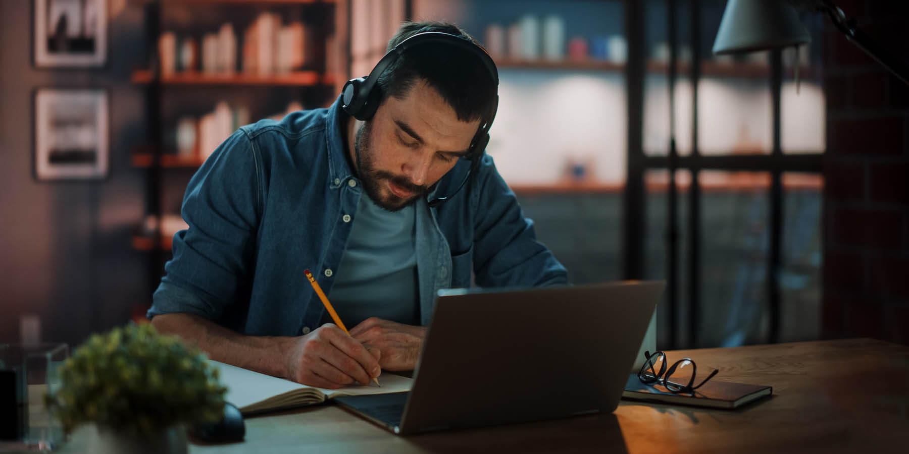Eine Person sitzt an einem Schreibtisch in einem schwach beleuchteten Raum und arbeitet an einem Laptop. Sie trägt Kopfhörer und hält einen Stift in der Hand. Auf dem Schreibtisch steht eine kleine Topfpflanze, und eine Brille liegt daneben. Im Hintergrund sind Bücherregale mit vielen Büchern zu sehen, und eine Lampe beleuchtet den Arbeitsbereich. Das Bild könnte mit Themen wie Fernarbeit, Studium, Homeoffice oder Konzentration in einer ruhigen Umgebung in Verbindung stehen.