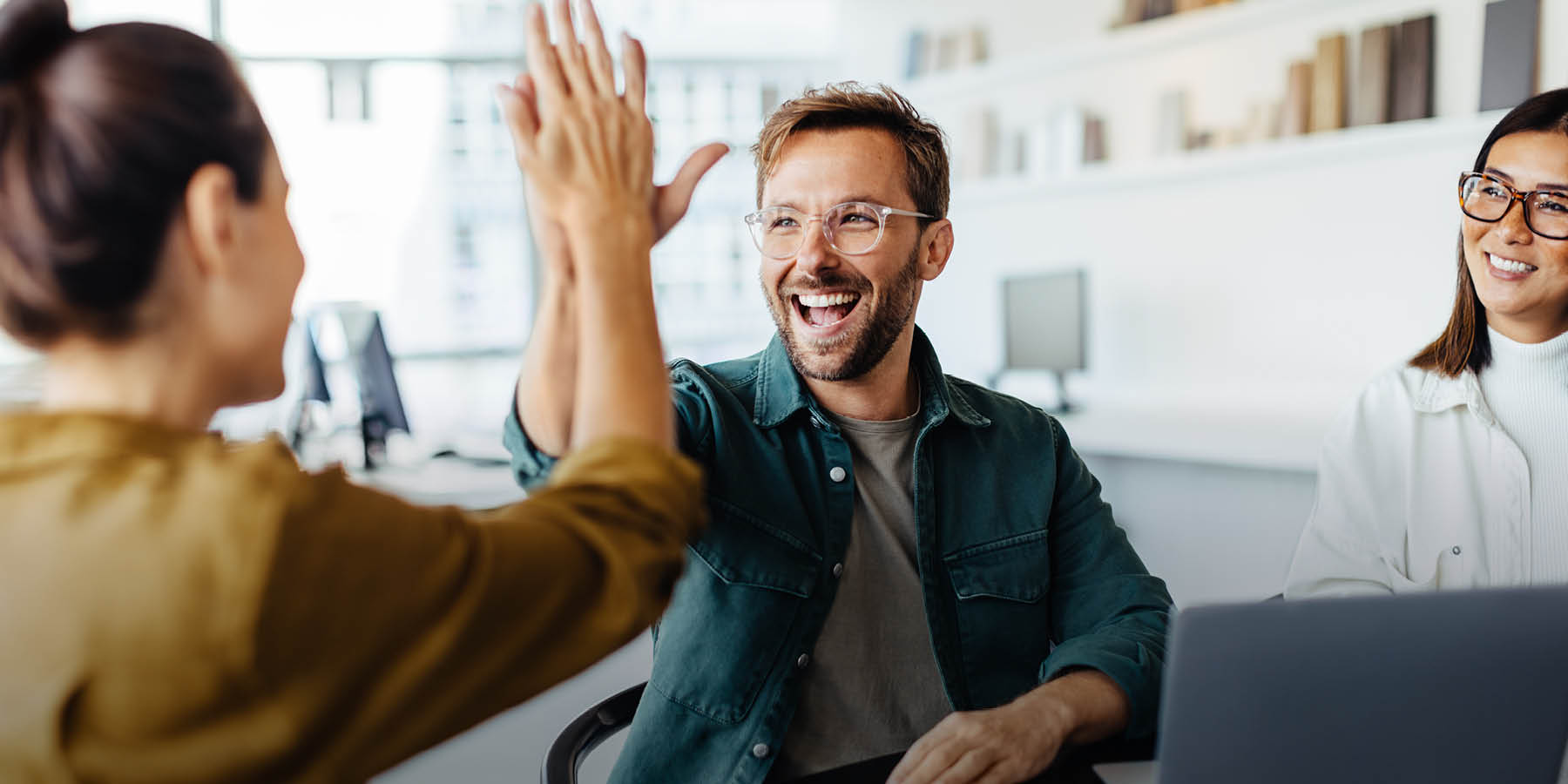 Drei Personen sitzen an einem Tisch in einem Büro und haben einen Laptop vor sich. Zwei Personen davon geben sich einen Handschlag mit erhobenen Händen und scheinen sich über einen Erfolg zu freuen.