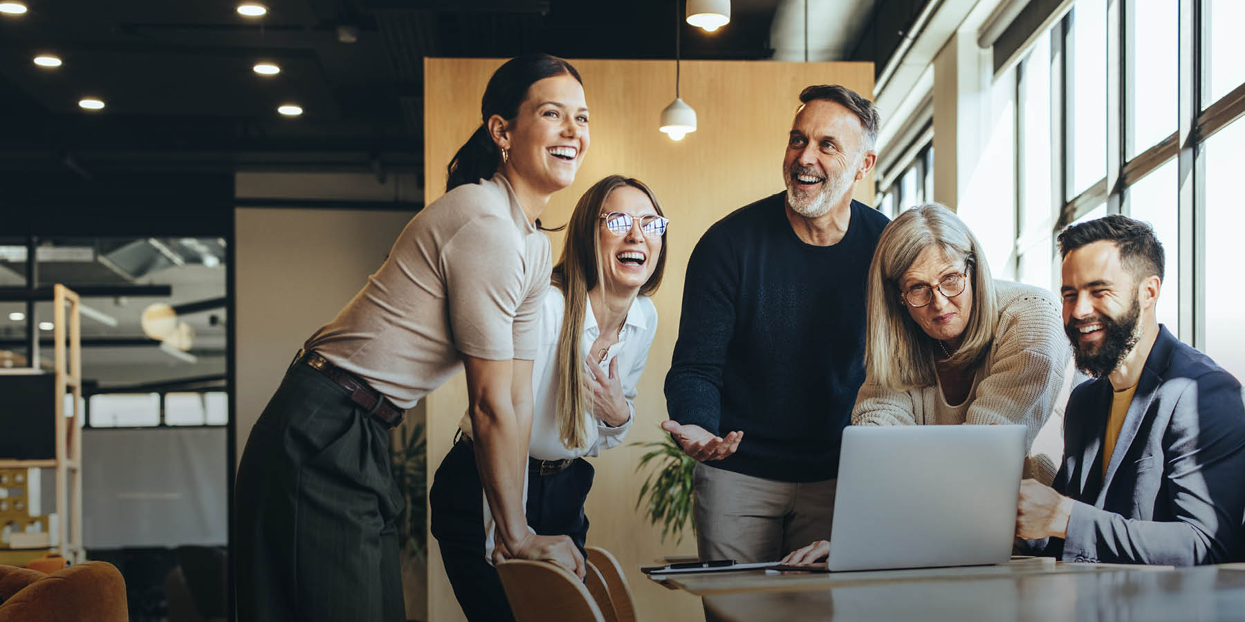 Eine Gruppe von Personen unterschiedlichen Alters stehen oder sitzen in einer modernen Büroumgebung an einem Tisch und lachen miteinander. Sie sind einem aufgeklappten Laptop zugewandt.