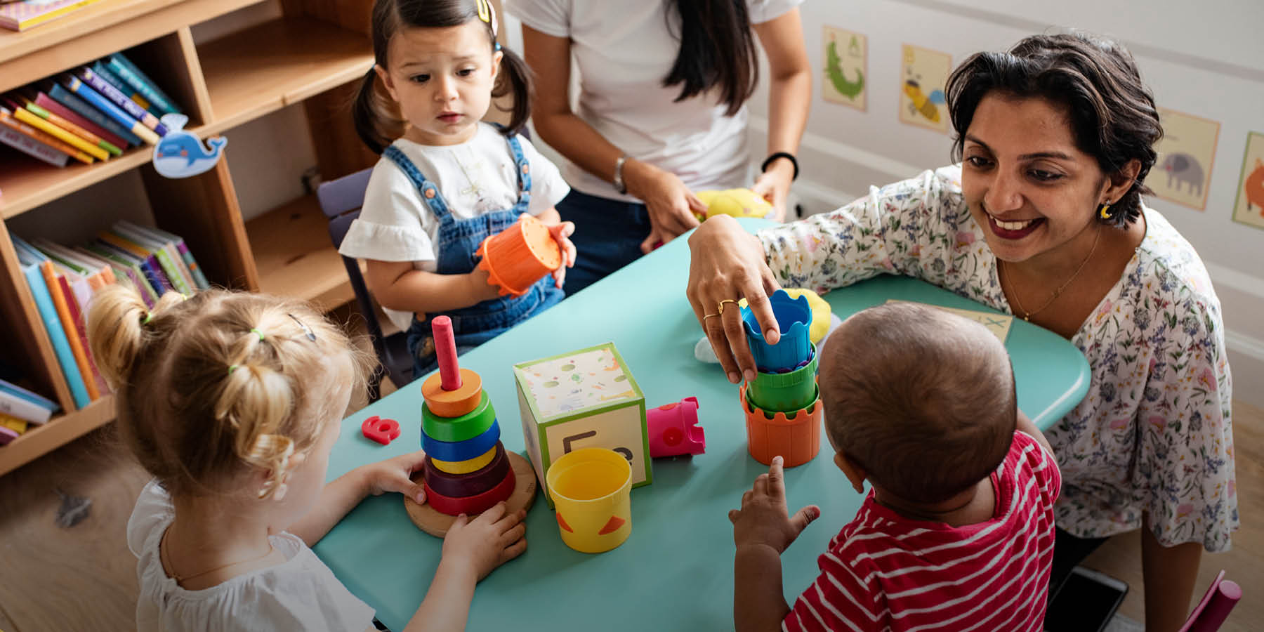 Eine Frau mittleren Alters kniet an einem Tisch und spielt mit drei Kindern im Kindergartenalter. Auf dem türkisfarbenen Tisch liegen verschiedene bunte Spielutensilien. An der Ecke des Tisches kann man noch einen Teil einer jüngeren Erzieherin erahnen.