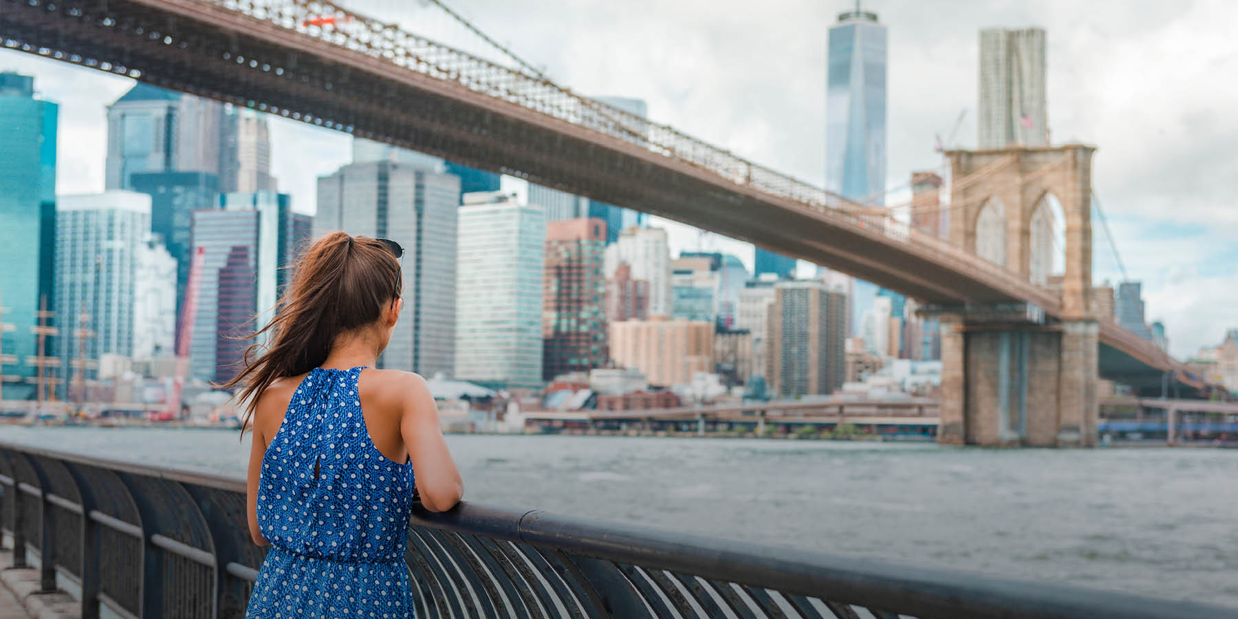 Eine junge Frau in einem blau gepunkteten Sommerkleid mit einem langen Pferdeschwanz und eine Sonnenbrille im Haar steht am Ufer des East River und schaut auf die Brooklyn Bridge und das dahinterliegende Brooklyn. Der Himmel ist bedeckt.