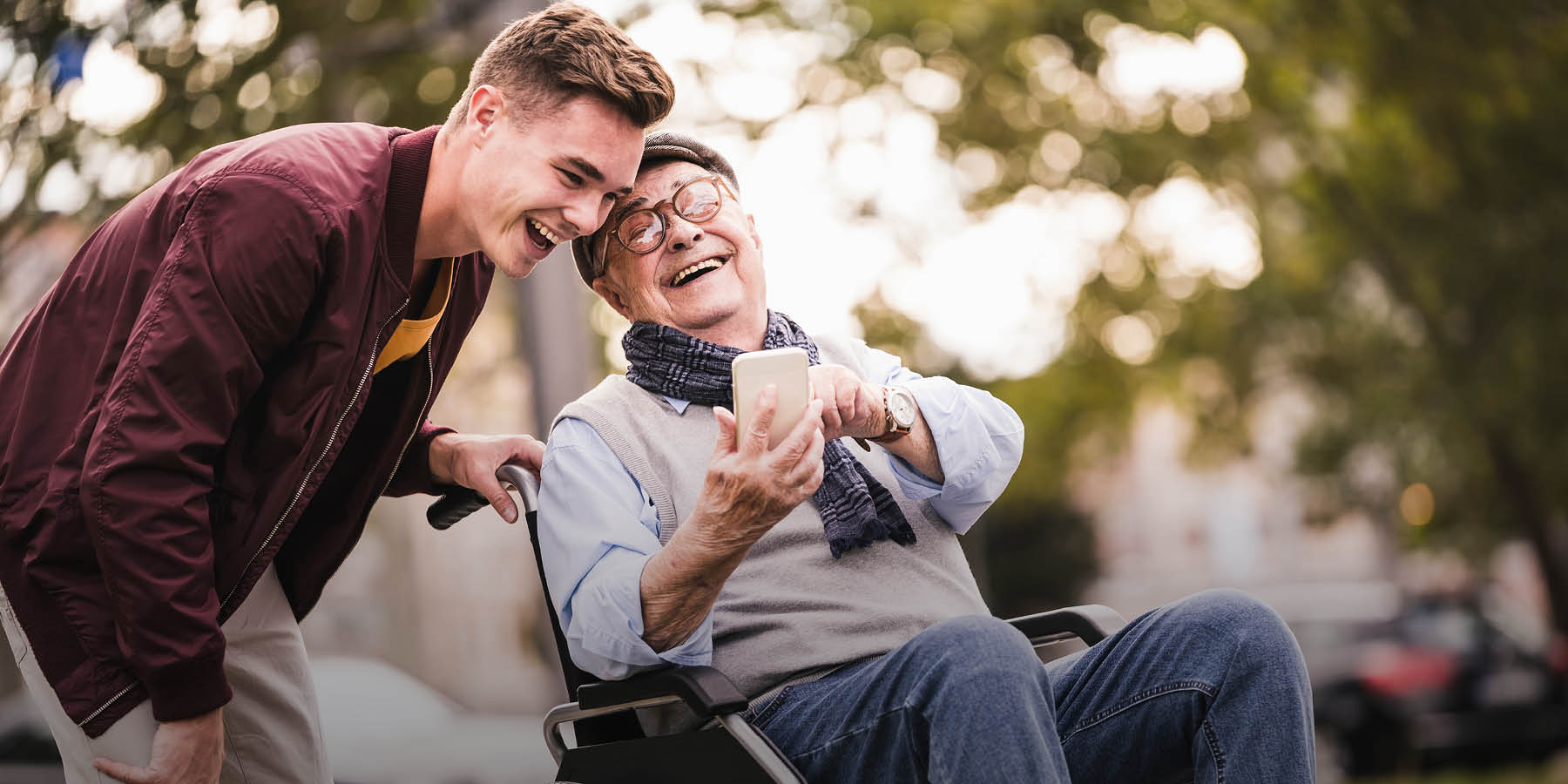 Ein älterer Mann mit Batschkappe und Brille, der in einem Rollstuhl sitzt, zeigt einem jüngeren Mann, der ihn schiebt, etwas auf seinem Smartphone. Beide Männer lachen. Die Stimmung ist herbstlich.