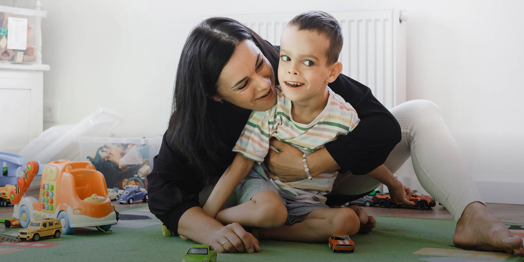 Eine jüngere Frau hält einen Jungen mit körperlicher Behinderung im Arm und albert mit ihm herum. Sie sitzen dabei auf einem Spielteppich, neben ihnen liegen verschiedene Spielzeugautos.