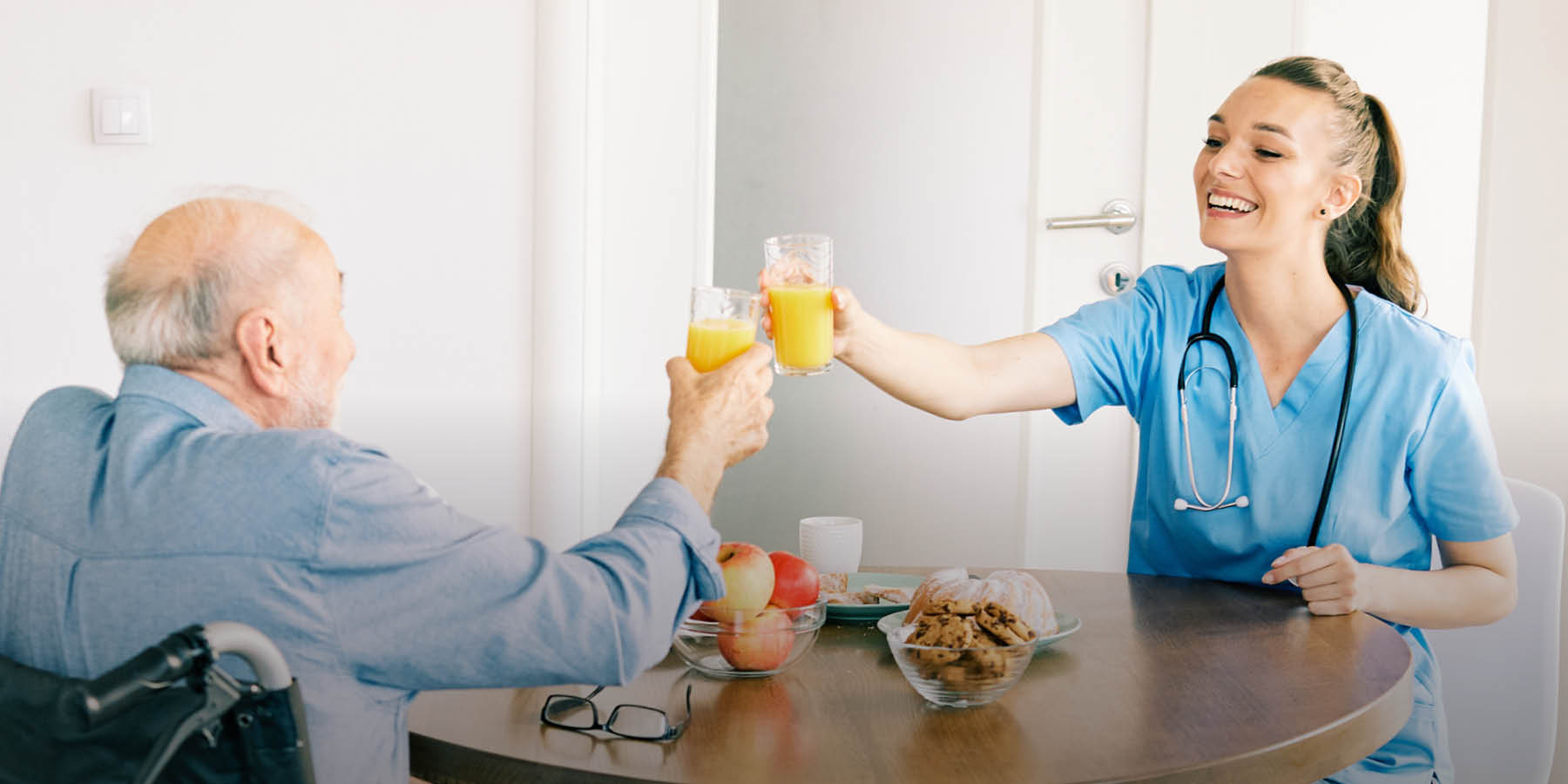 Ein älterer Mann im Rollstuhl und eine junge Pflegerin mit Stethoskop sitzen an einem runden Eichentisch und prosten sich mit Orangensaft zu. Auf dem Tisch stehen Glasschalen mit Keksen und Äpfeln.