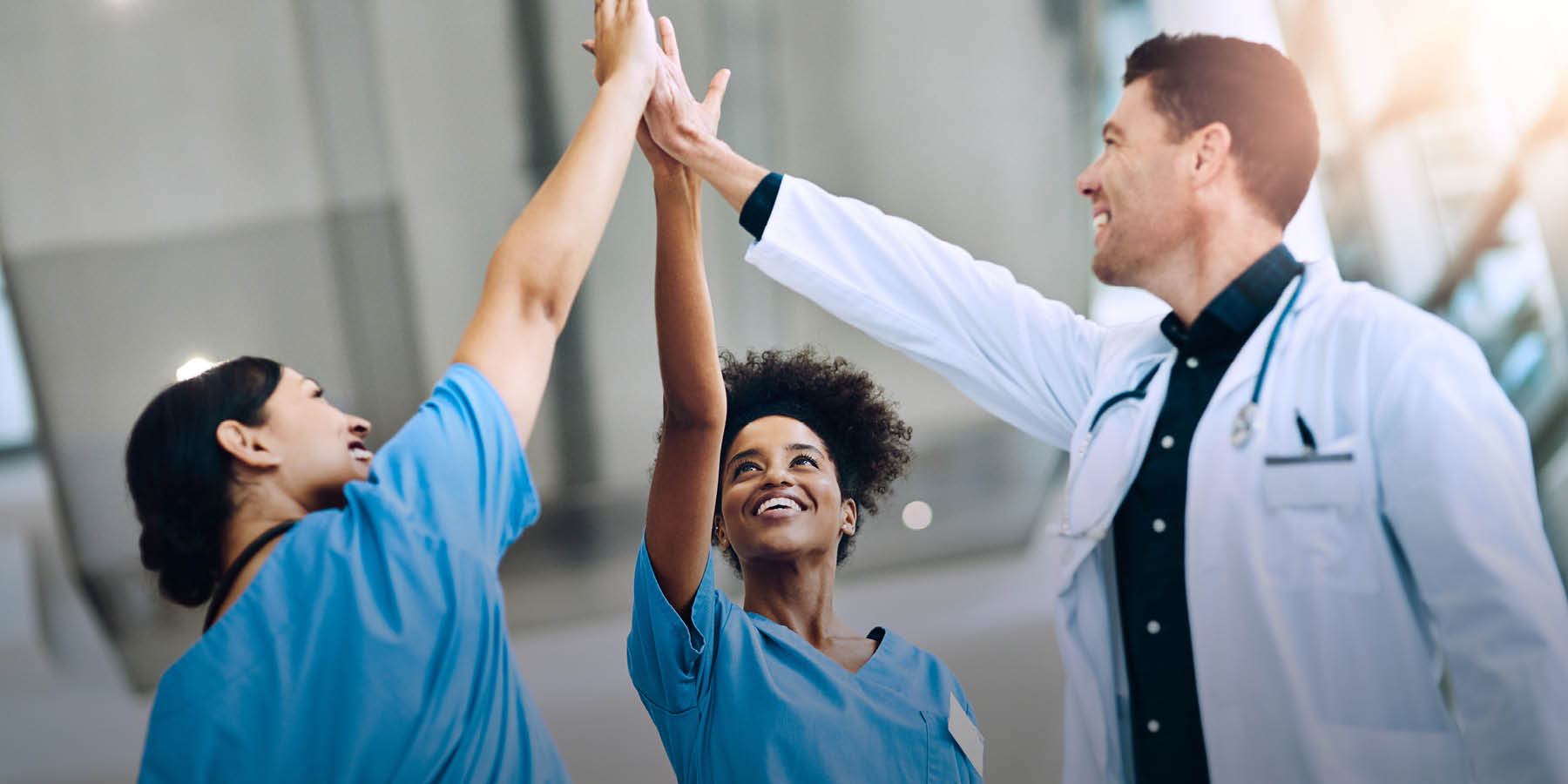 Zwei Pflegefachfrauen und ein Arzt machen eine High Five über ihren Köpfen. Das Bild ist leicht von unten aufgenommen.