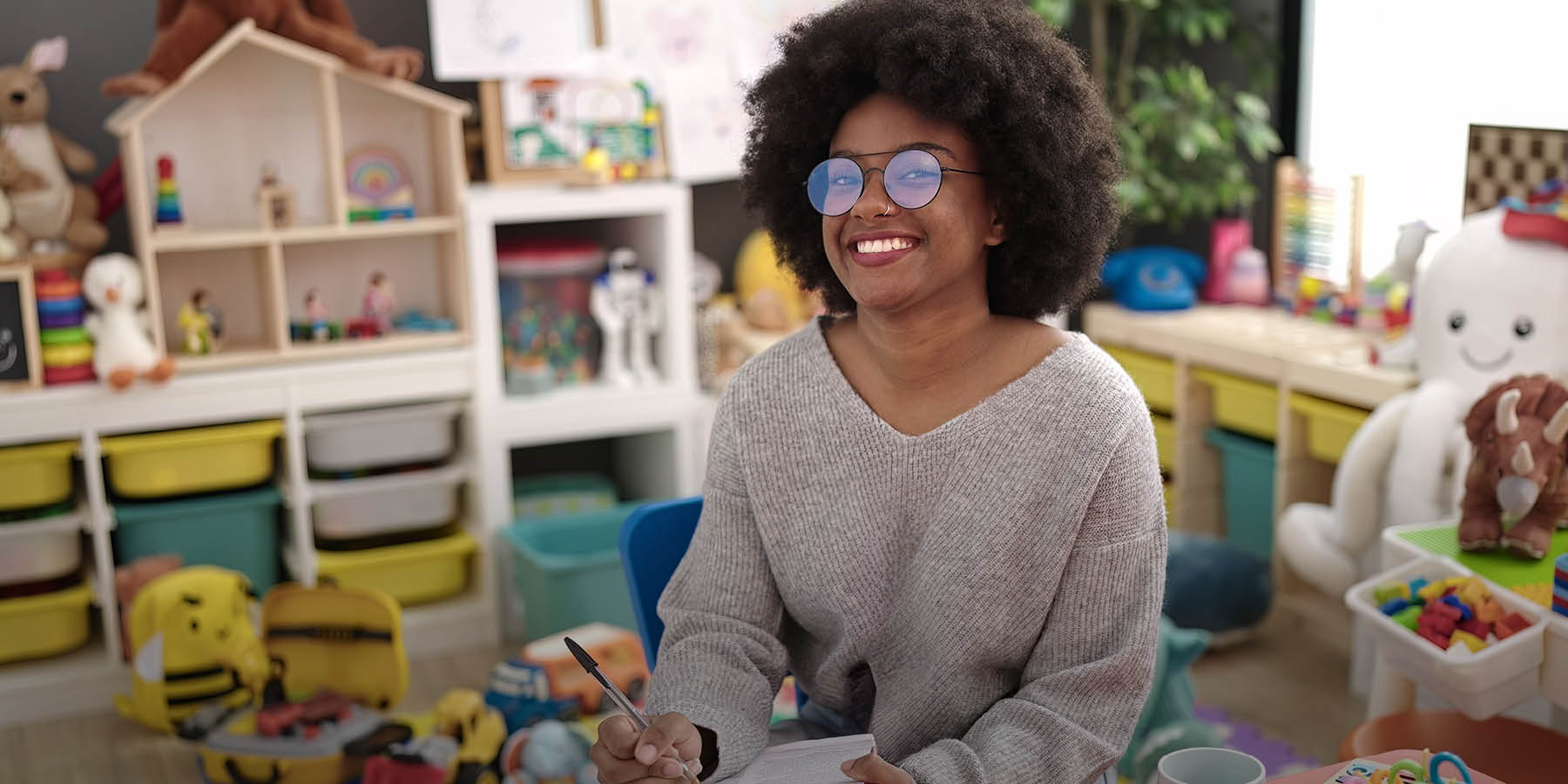 Eine junge, dunkelhäutige Frau mit dunklen Locken und Brille sitzt auf einem Stuhl. Sie hat ein Notizbuch und einen Stift auf ihrem Schoß liegen. Im Hintergrund erkennt man, dass die Szene in einem Kindergarten aufgenommen ist, da viele Kinderspielzeuge in den Schränken liegen, wie beispielsweise ein Puppenhaus.