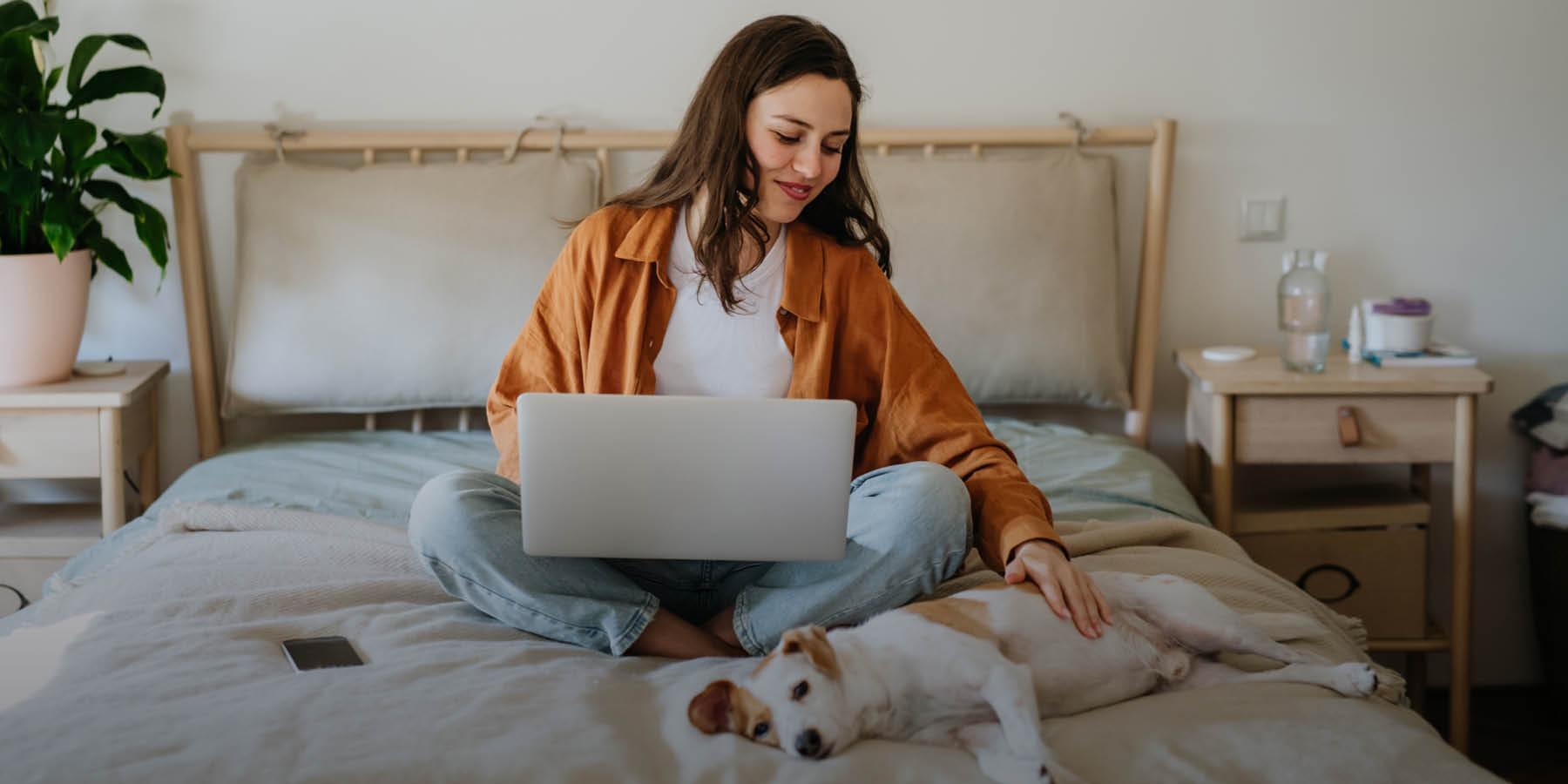 Eine Frau sitzt auf einem Bett und arbeitet an einem Laptop. Sie trägt eine braune Jacke über einem weißen T-Shirt und blaue Jeans. Neben ihr liegt ein kleiner Hund auf dem Bett, den sie streichelt. Das Zimmer ist hell und freundlich eingerichtet, mit einem Nachttisch, auf dem verschiedene Gegenstände wie eine Flasche und eine Pflanze stehen.