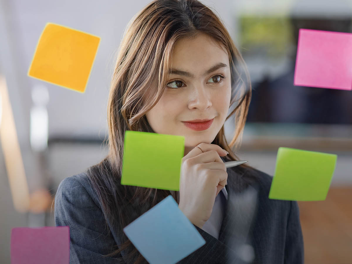 Eine junge Frau steht vor einer Glaswand auf der bunte Post-Its kleben. Sie hält einen Stift in der Hand, während sie diese ans Kinn hält und schaut nachdenklich. Der Hintergrund ist unscharf.