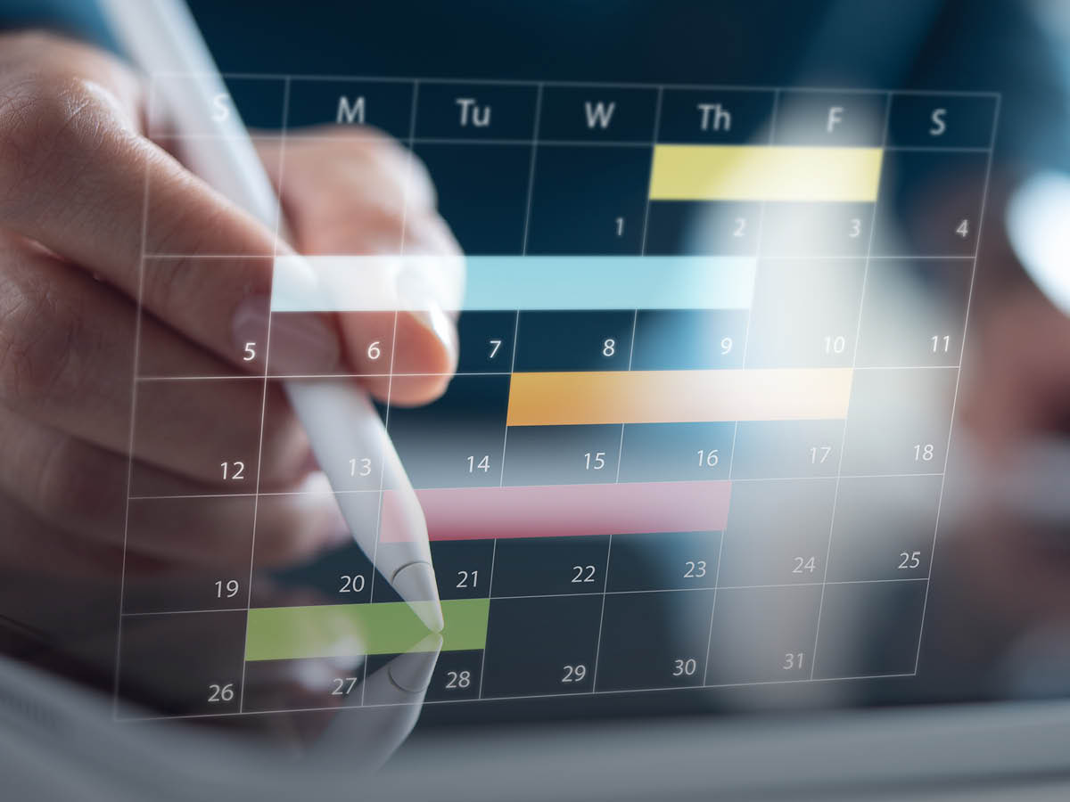 Eine Hand mit Eingabestift bedient einen digitalen Monatskalender auf einem Tablet. Mehrere farbige Balken markieren Aufgaben, Termine und Projektphasen über verschiedene Tage – Symbol für digitale Zeitplanung und effizientes Projektmanagement.