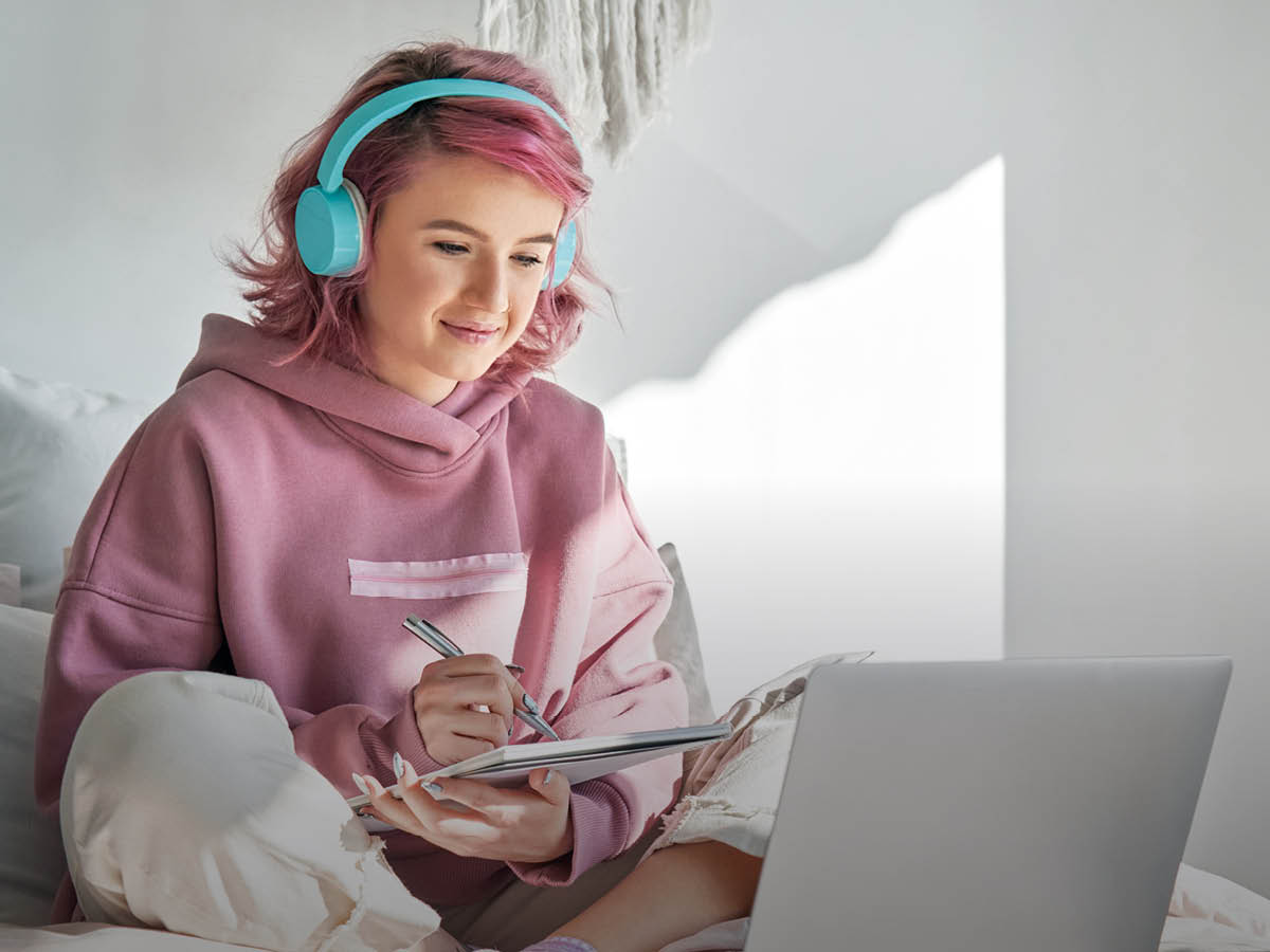 Eine junge Frau mit rosa Haaren, rosa Kapuzenpullover und türkisfarbenen Kopfhörern sitzt im Schneidersitz auf einem Bett vor einem aufgeklappten Laptop und hält einen Notizblock und einen Stift in den Händen.
