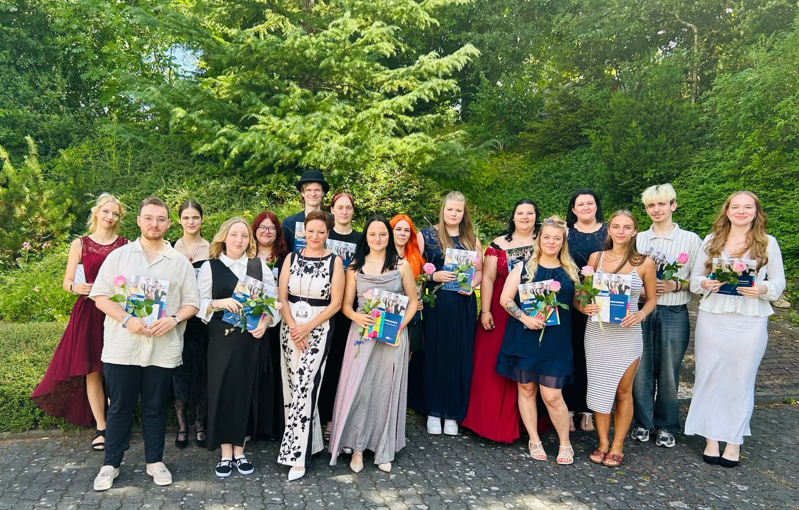 Eine Gruppe festlich gekleideter Absolvent*innen steht mit Rosen und Unterlagen für ein gemeinsames Erinnerungsfoto im Grünen.