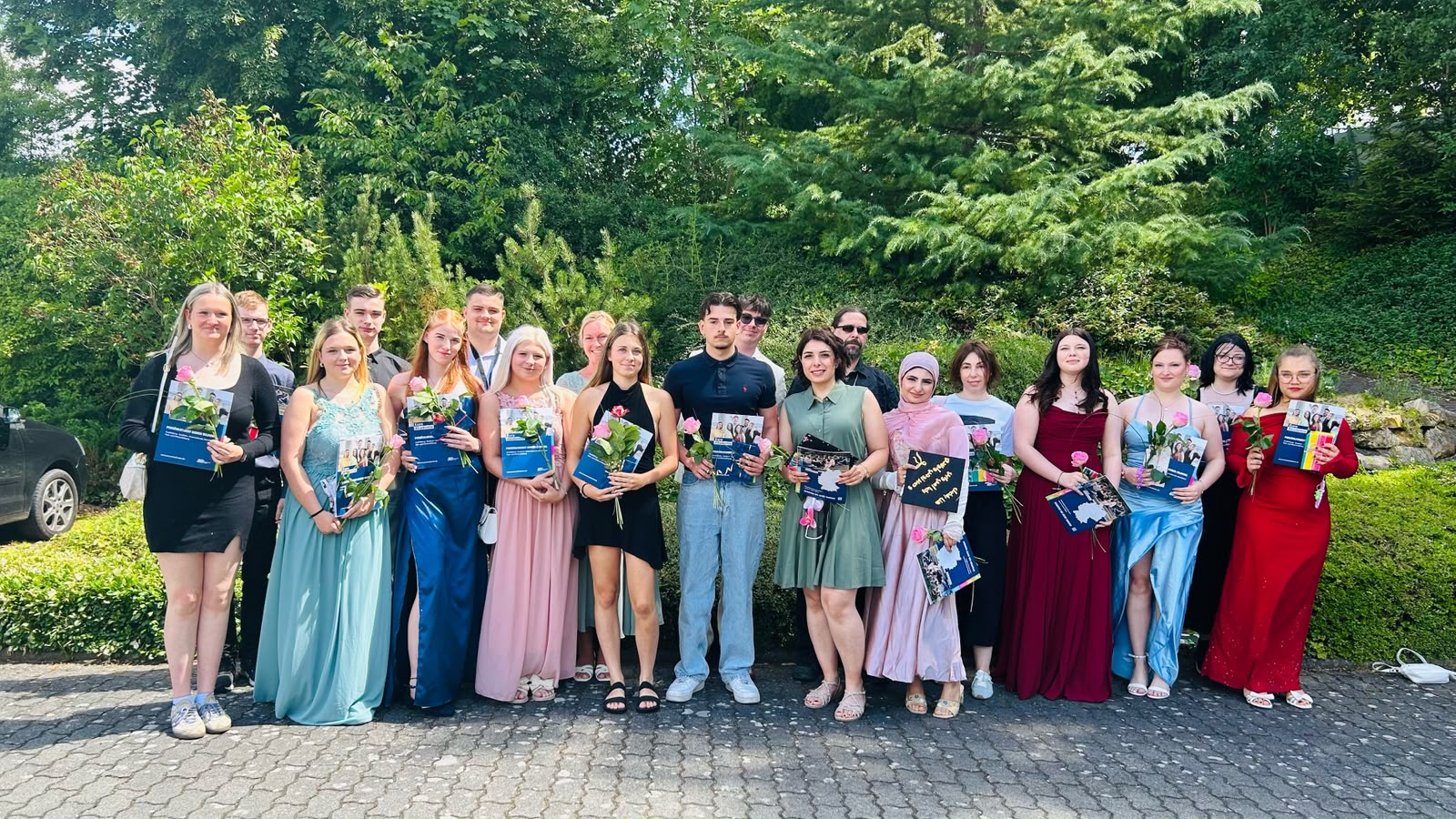 Eine große Gruppe Absolvent*innen steht in festlicher Kleidung mit Rosen und Abschlussmappen für ein Gruppenfoto im Freien.