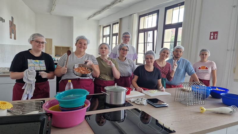 Auszubildende stehen in einer hellen Küche an einer Arbeitsfläche. Sie tragen Haarnetze und Schürzen. Auf dem Tisch liegen verschiedene Küchenutensilien, darunter Schüsseln, ein Topf, ein Schneidebrett und ein Metallgestell. Eine Person hält eine Schüssel mit geschnittenen Zutaten, andere schneiden oder bereiten Lebensmittel vor. Die Fenster im Hintergrund lassen viel Tageslicht herein.