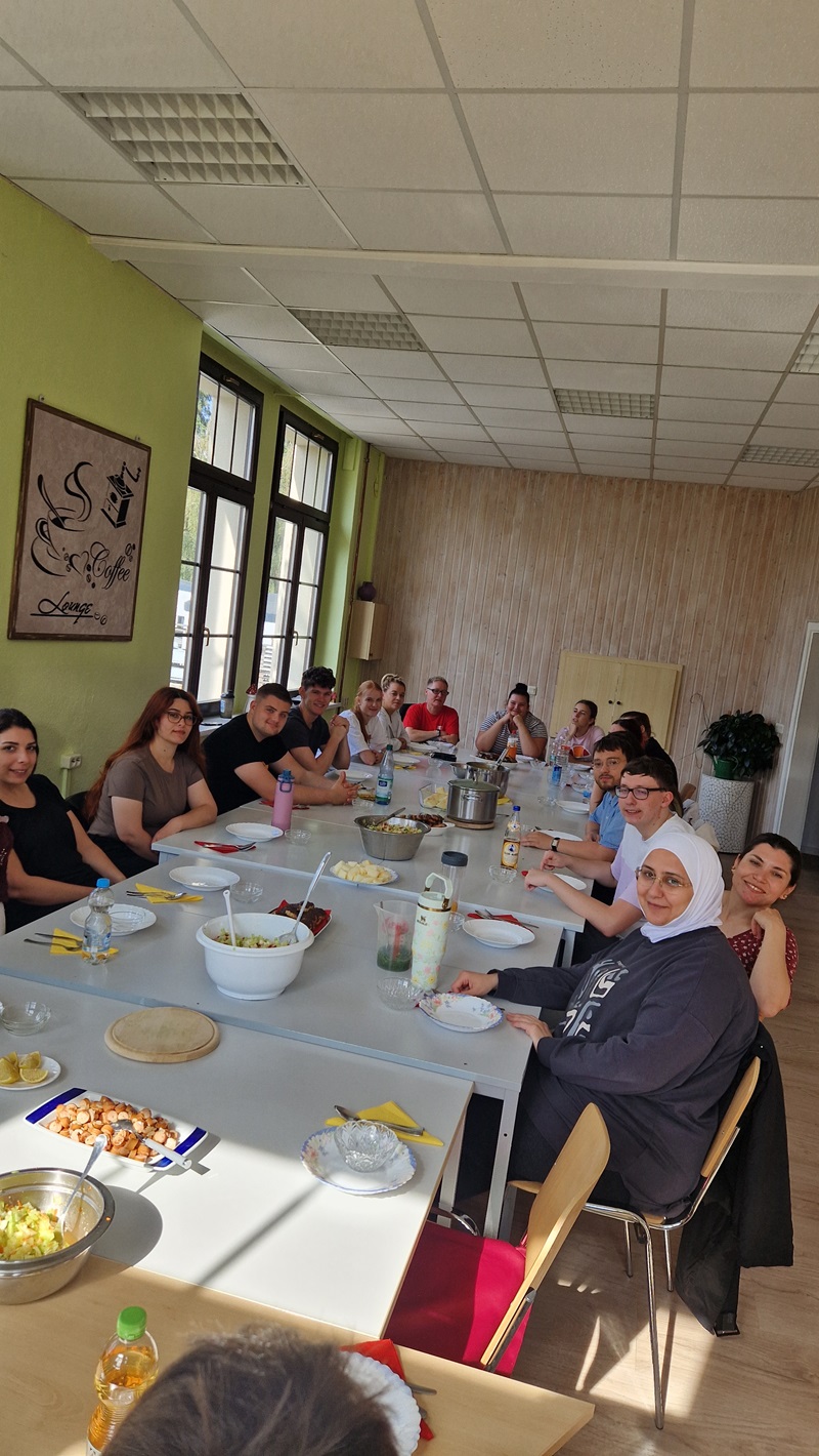 Eine Gruppe von Azubis sitzt an einem langen Tisch in einem hellen Raum mit großen Fenstern. Auf dem Tisch stehen verschiedene Schüsseln mit Salat, Brot, Getränkeflaschen und weiteren Speisen. Die Personen schauen in Richtung Kamera, einige lächeln. Die Wände sind hellgrün und beige, an einer Wand hängt ein großes gerahmtes Bild mit kalligrafischen Elementen. Im Hintergrund steht eine große Pflanze in einem weißen Topf.