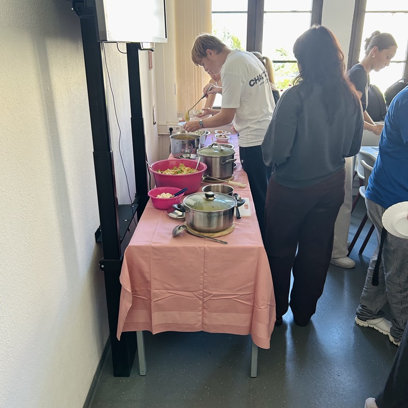 Ein langer Tisch ist mit einem rosa Tischtuch bedeckt und steht an einer Wand. Darauf stehen mehrere Edelstahltöpfe mit Deckeln, eine große Metallschüssel mit Salat und zwei pinkfarbene Schüsseln. Mehrere Auszubildende stehen seitlich am Tisch und bedienen sich am Buffet. Tageslicht fällt durch große Fenster auf die Szene.