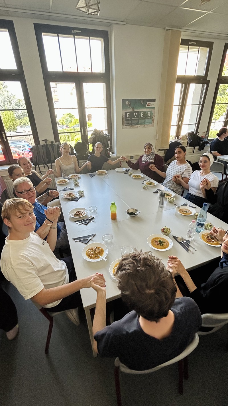 Die Gruppe Auszubildender sitzt an einem langen, weißen Tisch in einem hellen Raum mit großen Fenstern. Auf dem Tisch stehen Teller mit Essen, Gläser und eine Flasche mit grünem Deckel. Die Personen halten sich an den Händen und schauen in Richtung Kamera. An der Wand hängt ein Poster mit einem Landschaftsmotiv. Draußen sind Bäume und ein Gebäude zu sehen.