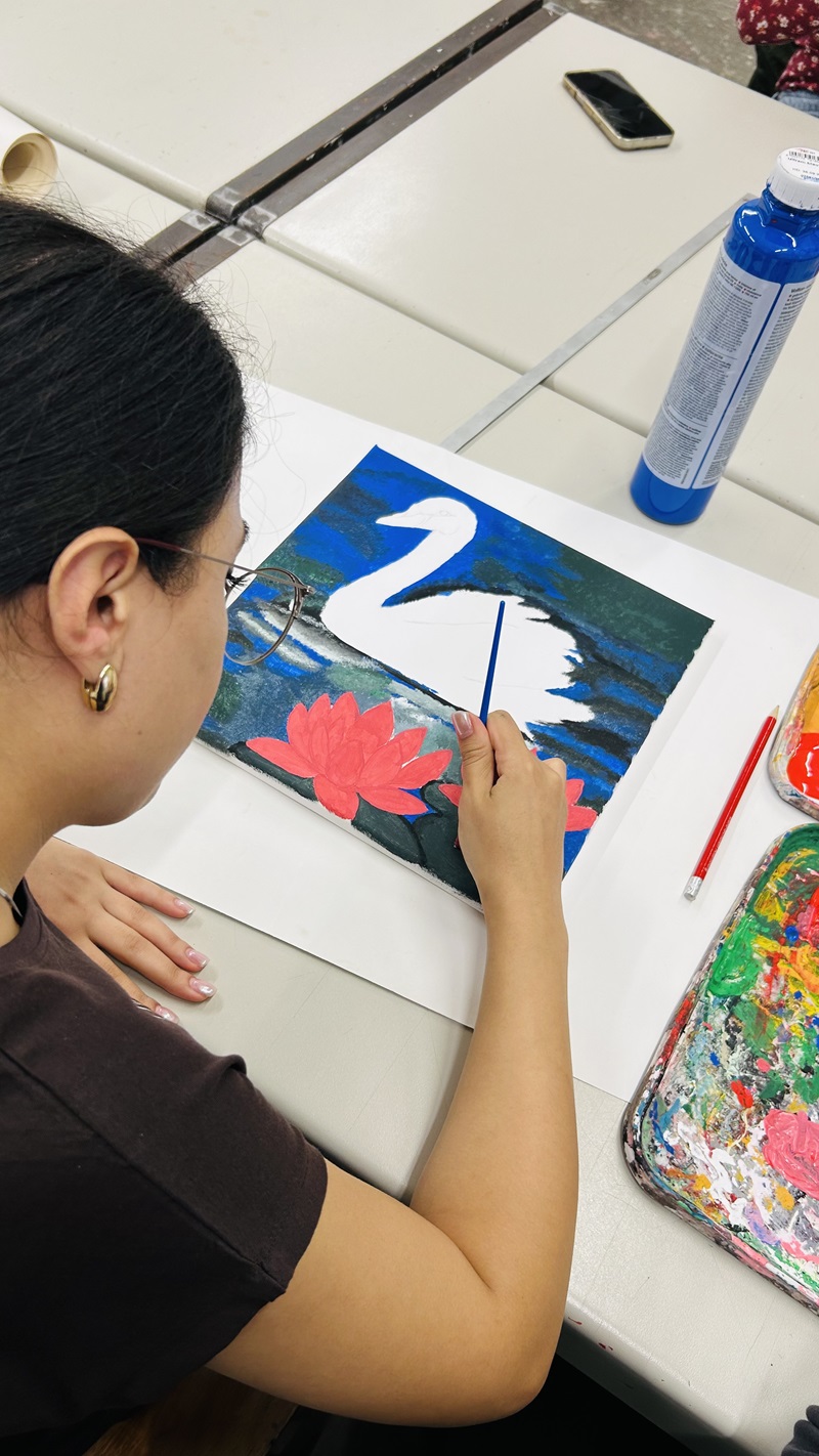 Eine Auszubildende mit dunklen Haaren und goldenen Ohrringen sitzt an einem Tisch und malt mit einem feinen Pinsel auf einem Blatt Papier. Das Motiv zeigt einen weißen Schwan auf einem blauen Hintergrund mit roten Seerosen im Vordergrund. Neben dem Blatt liegen ein roter Bleistift, eine Tube blaue Farbe und eine Farbpalette mit verschiedenen Farbresten.