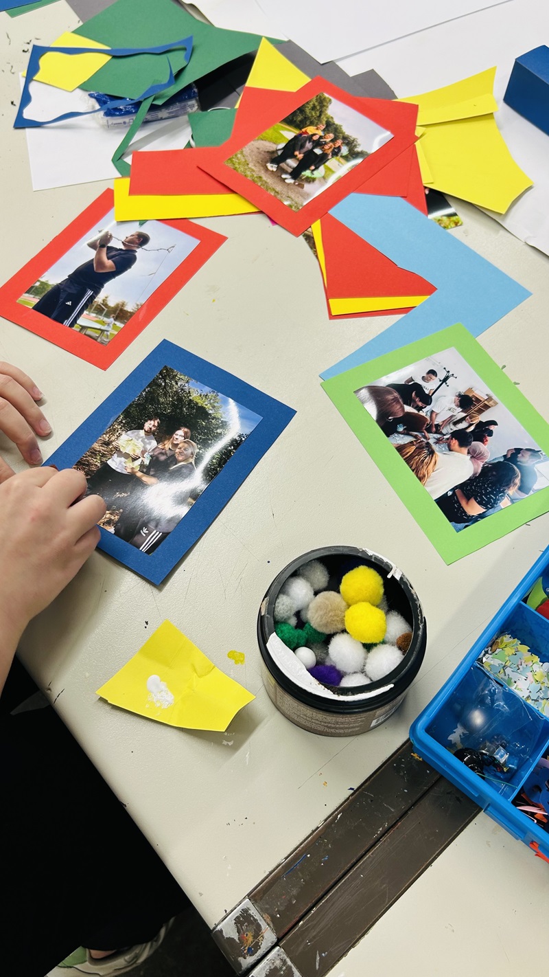 Auf einem Tisch liegen mehrere farbige Papierstücke, Fotos mit bunten Rahmen und Bastelmaterialien wie kleine, flauschige Bälle. Eine Hand hält ein Foto mit blauem Rahmen, auf dem eine Gruppe von Menschen im Freien zu sehen ist. Daneben stehen eine Dose mit bunten Pompons und eine blaue Box mit weiteren Bastelutensilien. Die Szene zeigt eine Bastelarbeit mit Fotos und Dekoration.