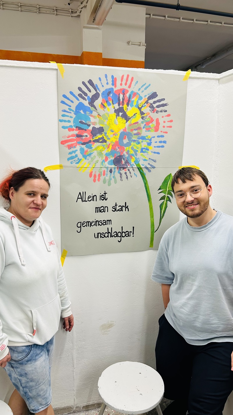 Zwei Auszubildende stehen nebeneinander vor einer weißen Wand. Zwischen ihnen hängt ein großes Plakat, das eine bunte Blume zeigt. Die Blütenblätter bestehen aus vielen unterschiedlich farbigen Handabdrücken. Der Stiel und die Blätter sind grün gemalt. Auf dem Plakat steht in klarer Schrift: 'Allein ist man stark, gemeinsam unschlagbar!'. Die Szene wirkt freundlich und gemeinschaftlich.