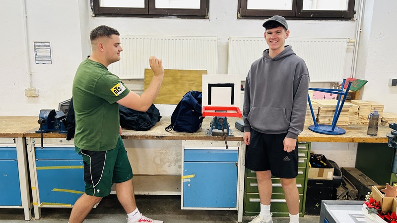 Zwei männliche Azubis stehen in einer Werkstatt vor einer Werkbank. Einer trägt ein grünes T-Shirt und Shorts, der andere einen grauen Kapuzenpullover und eine schwarze Kappe. Auf der Werkbank liegen verschiedene Werkzeuge und Materialien, darunter ein roter Rahmen und ein blauer Hocker. Im Hintergrund sind weiße Heizkörper und Fenster zu sehen.