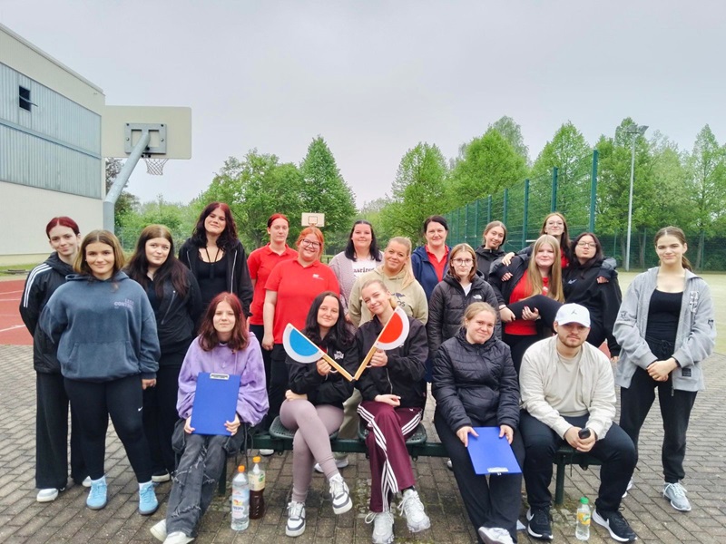 Die Gruppe der Auszubildenden der Euro Akademie Altenburg steht und sitzt auf einer Bank im Freien vor einem Sportplatz. Im Hintergrund sind ein Basketballkorb, ein Zaun und Bäume zu sehen. Einige Personen tragen rote Oberteile, andere dunkle oder helle Sportkleidung. Zwei Personen in der Mitte halten bunte Schläger in den Händen. Auf dem Boden stehen mehrere Getränkeflaschen. Die Stimmung wirkt entspannt.