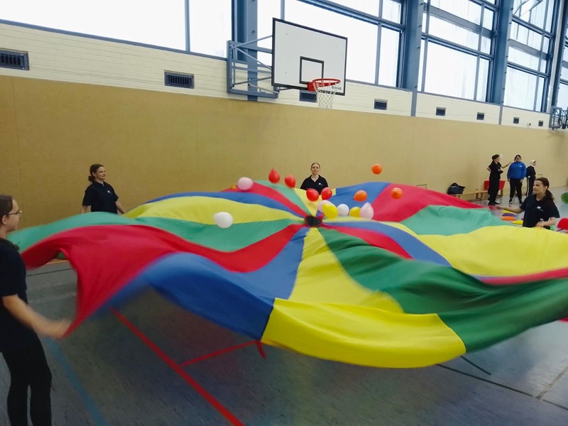 Mehrere Auszubildende stehen in einer Sporthalle und halten gemeinsam ein großes, buntes Schwungtuch, das in verschiedene farbige Segmente unterteilt ist. Auf dem Schwungtuch liegen mehrere bunte Bälle, die durch die Bewegung des Tuchs in die Luft geworfen werden. Im Hintergrund sind eine Basketballkorbanlage, hohe Fenster und eine beige Wand zu sehen.
