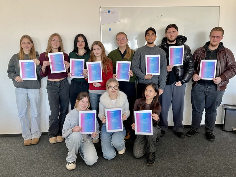 Die Gruppe von elf Teilnehmenden der Euro Akademie Aschaffenburg stehen und knien teilweise vor einer weißen Wand mit einem Whiteboard. Alle halten jeweils eine Urkunde mit einem Farbverlauf von Blau zu Pink, die den Europass "Mobilität" des Erasmus+-Programms symbolisiert in den Händen. Die Personen tragen unterschiedliche, legere, moderne Kleidung, darunter Pullover, Jacken und Jeans. Die Szene zeigt die feierliche Übergabe der Europässe an die Teilnehmenden, die sie freudig entgegen nehmen.