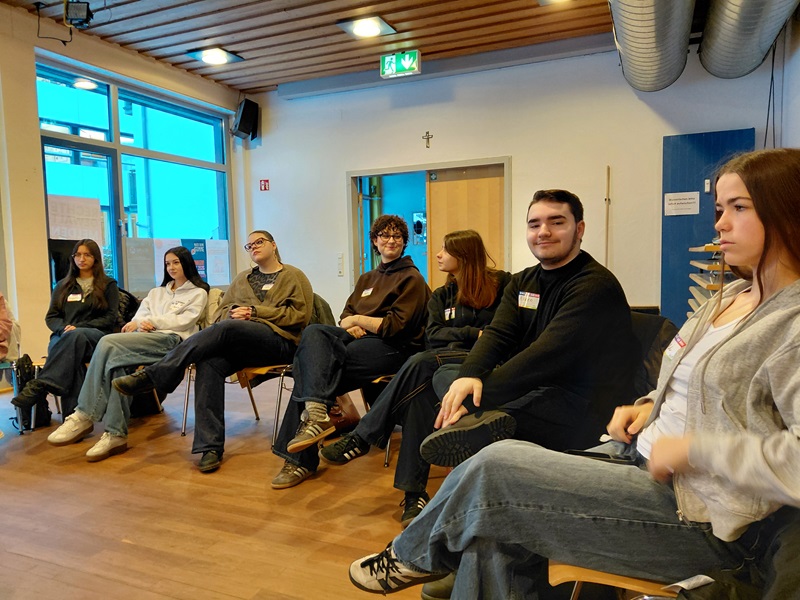 Mehrere junge Azubis sitzen in einem hellen Seminarraum in einem lockeren Stuhlkreis. Die Auszubildenden tragen Namensschilder und sind leger gekleidet. Im Hintergrund sind große Fenster, eine Holztür, eine blaue Wand und sichtbare Lüftungsrohre zu sehen. Die Szene entstand während des Medien-Workshops „Kommunalwahlen on Air“ mit Auszubildenden zu Fremdsprachenkorrespondent*innen der Euro Akademie Aschaffenburg. Die Atmosphäre wirkt konzentriert und offen, die Teilnehmenden blicken aufmerksam in Richtung einer Person außerhalb des Bildausschnitts.