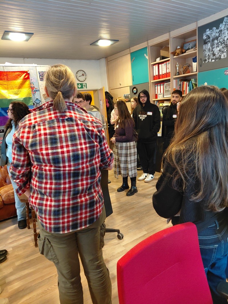 In einem hellen Seminarraum mit Holzfußboden und Regalen voller Ordner und Bücher stehen mehrere junge Schülerinnen und Schüler in lockerer Runde beisammen. Im Vordergrund ist eine Person mit kariertem Hemd von hinten zu sehen. Im Hintergrund hängen eine Regenbogenflagge und verschiedene Poster an der Wand. Die Teilnehmenden tragen Namensschilder und sind überwiegend leger gekleidet. Die Szene entstand während des Medien-Workshops „Kommunalwahlen on Air“ mit Auszubildenden zu Fremdsprachenkorrespondent*innen der Euro Akademie Aschaffenburg.