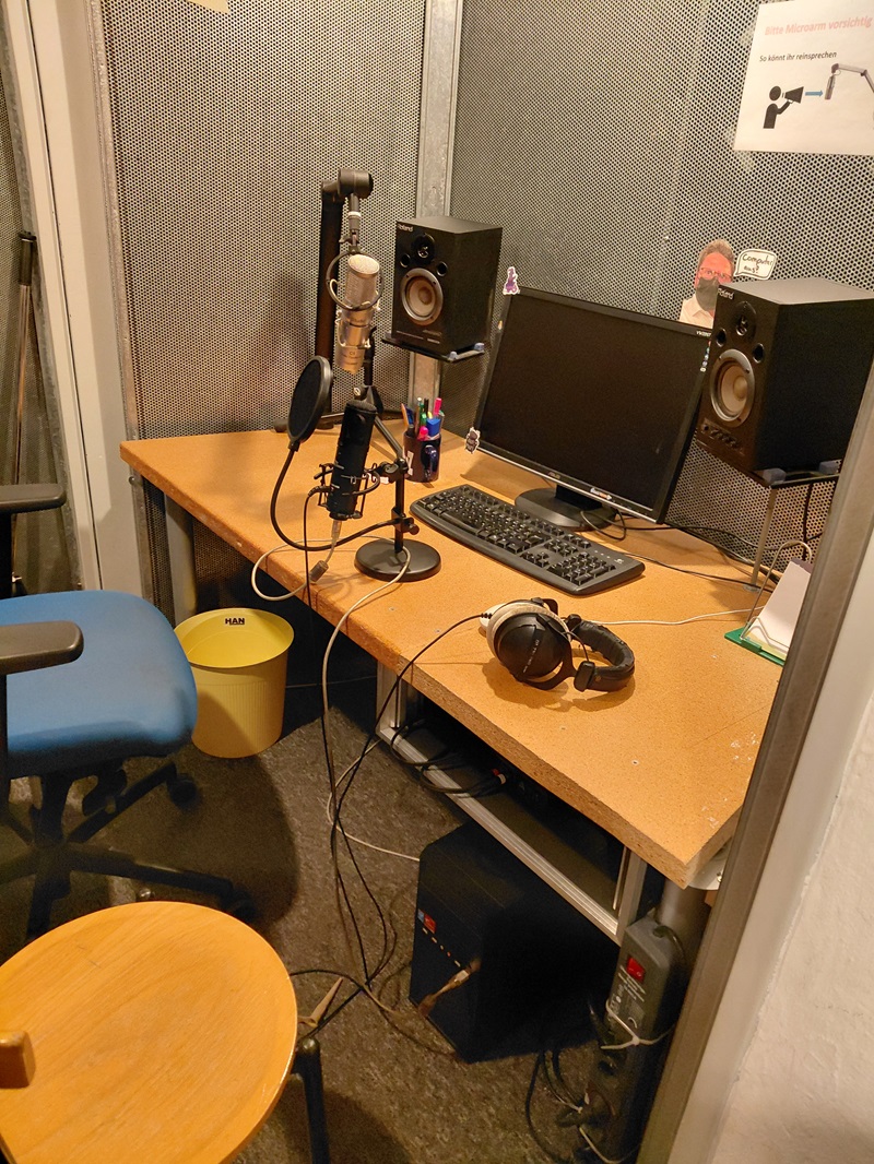 Tonstudio-Arbeitsplatz mit Mikrofon, Popfilter, Kopfhörer, Studiomonitoren, Computerbildschirm, Tastatur und Maus auf einem Holzschreibtisch. Zwei Stühle stehen nebeneinander, einer davon ist gepolstert und höhenverstellbar. An der Wand hängt ein Informationsblatt. Die Szene entstand während des Medien-Workshops „Kommunalwahlen on Air“ mit Auszubildenden zu Fremdsprachenkorrespondent*innen der Euro Akademie Aschaffenburg.