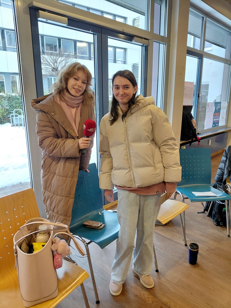Zwei weibliche Personen in Winterkleidung stehen drinnen vor einem großen Fenster. Draußen ist Schnee zu sehen. Eine Person hält ein rotes Mikrofon und trägt einen langen Daunenmantel, die andere eine weiße Daunenjacke und helle Jeans. Auf einem Stuhl in der Nähe steht eine beige Tasche mit einem Stofftier. Es scheint sich um einen Wartebereich mit Holzboden und blauen Stühlen zu handeln.