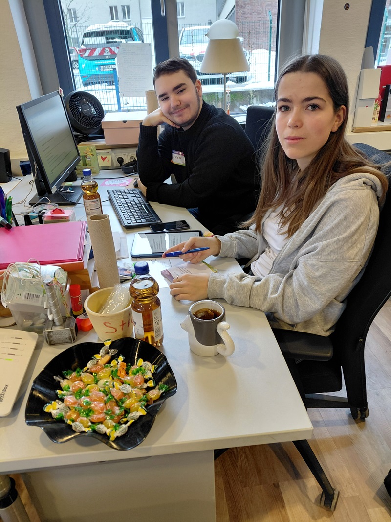 Zwei Auszubildende sitzen an einem Schreibtisch. Eine Person lächelt und arbeitet am Computer, während die andere mit einem Stift in der Hand in die Kamera schaut. Auf dem Schreibtisch stehen verschiedene Gegenstände, darunter eine schwarze Schale mit bunten Bonbons, eine Flasche, eine Tasse Tee und Büromaterial. Durch ein Fenster im Hintergrund sieht man draußen einige Gebäude und ein geparktes Auto.