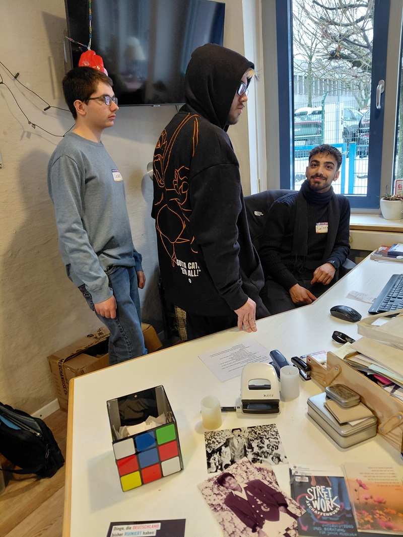 Drei Auszubildende befinden sich in einem Raum: eine Person steht und trägt eine Brille mit Namensschild, eine weitere Person steht ebenfalls in einem Kapuzenpullover, und eine dritte Person sitzt und lächelt leicht. Auf einem Schreibtisch im Vordergrund liegen verschiedene Gegenstände, darunter ein großer Zauberwürfel, ein Locher und einige Fotos. Im Hintergrund sind ein Fenster und ein Monitor zu sehen.