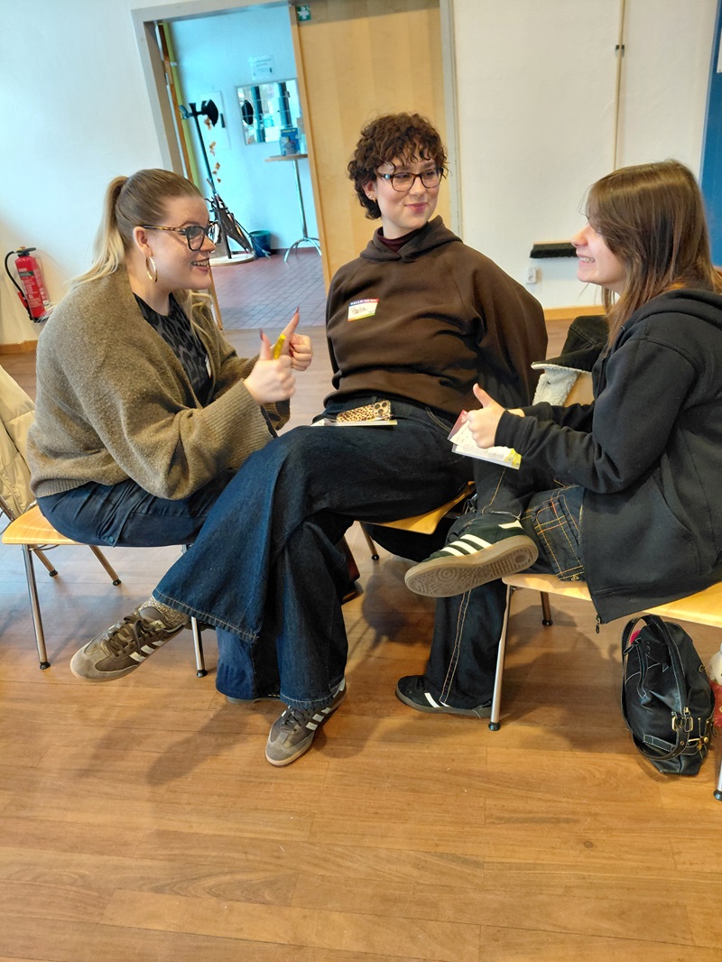 Drei Erwachsene sitzen in einem Raum auf Stühlen und unterhalten sich. Zwei von ihnen zeigen den Daumen nach oben, eine lächelt. Sie sind leger gekleidet und scheinen sich angeregt zu unterhalten. Der Raum hat einen Holzboden, und im Hintergrund sind einige Geräte und ein Feuerlöscher zu sehen.