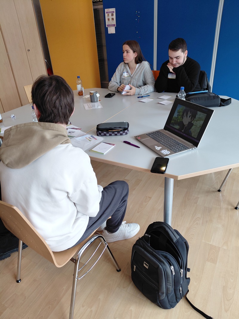 Schülerinnen und Schüler sitzen an einem Tisch in einem Raum mit leuchtend gelben und blauen Trennwänden. Sie unterhalten sich; auf dem Tisch liegen Notizen, Flaschen und ein Laptop. Auf dem Laptop-Bildschirm ist eine Anime-Szene zu sehen. Rucksäcke stehen neben den Stühlen auf dem Boden.