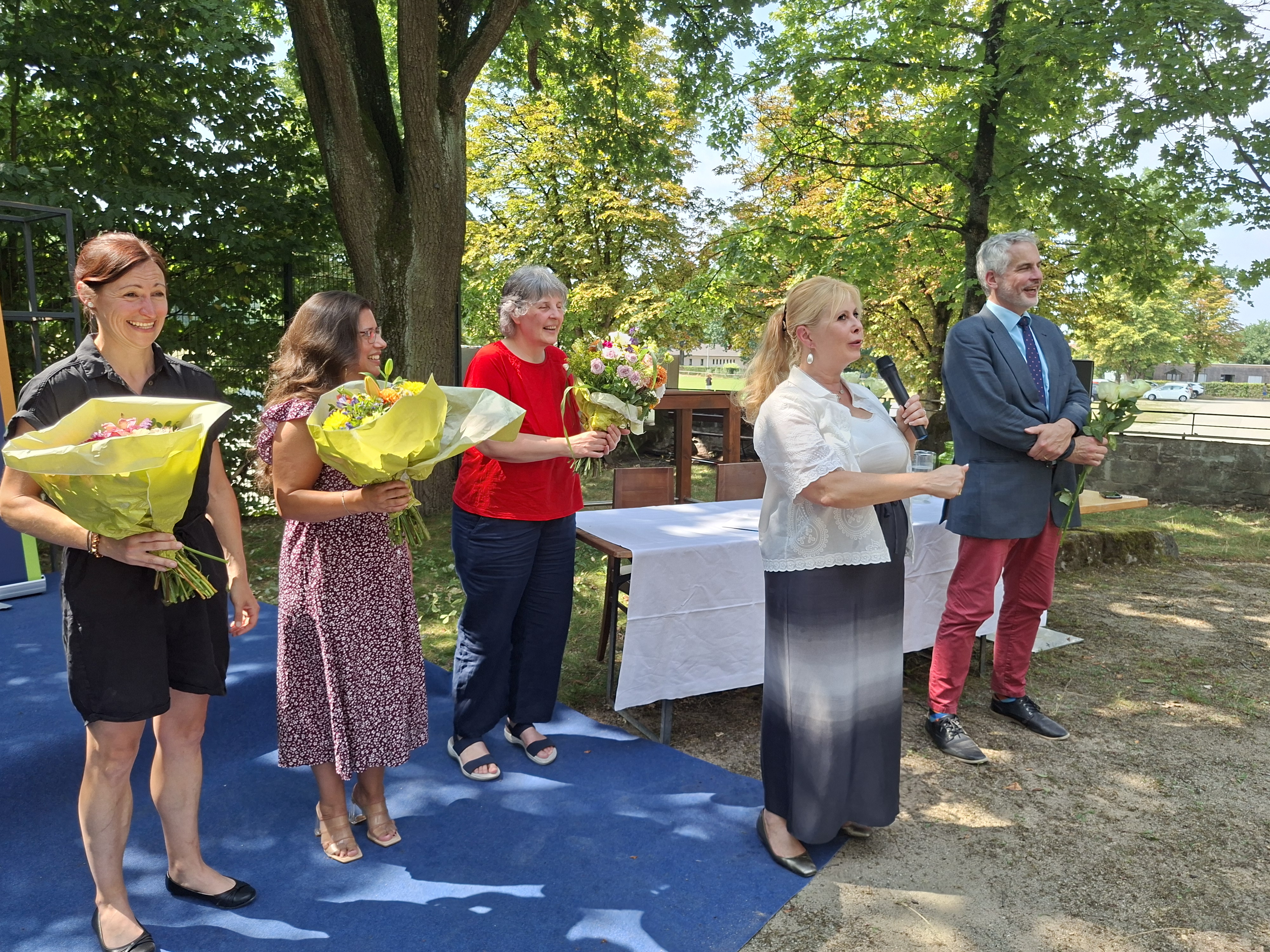 Vor einer kleinen Bühne im Freien stehen drei Frauen mit Blumensträußen. Rechts vorne stehen ein Mann mit Rosen in den Händen und eine weitere Frau mit Mikrofon.