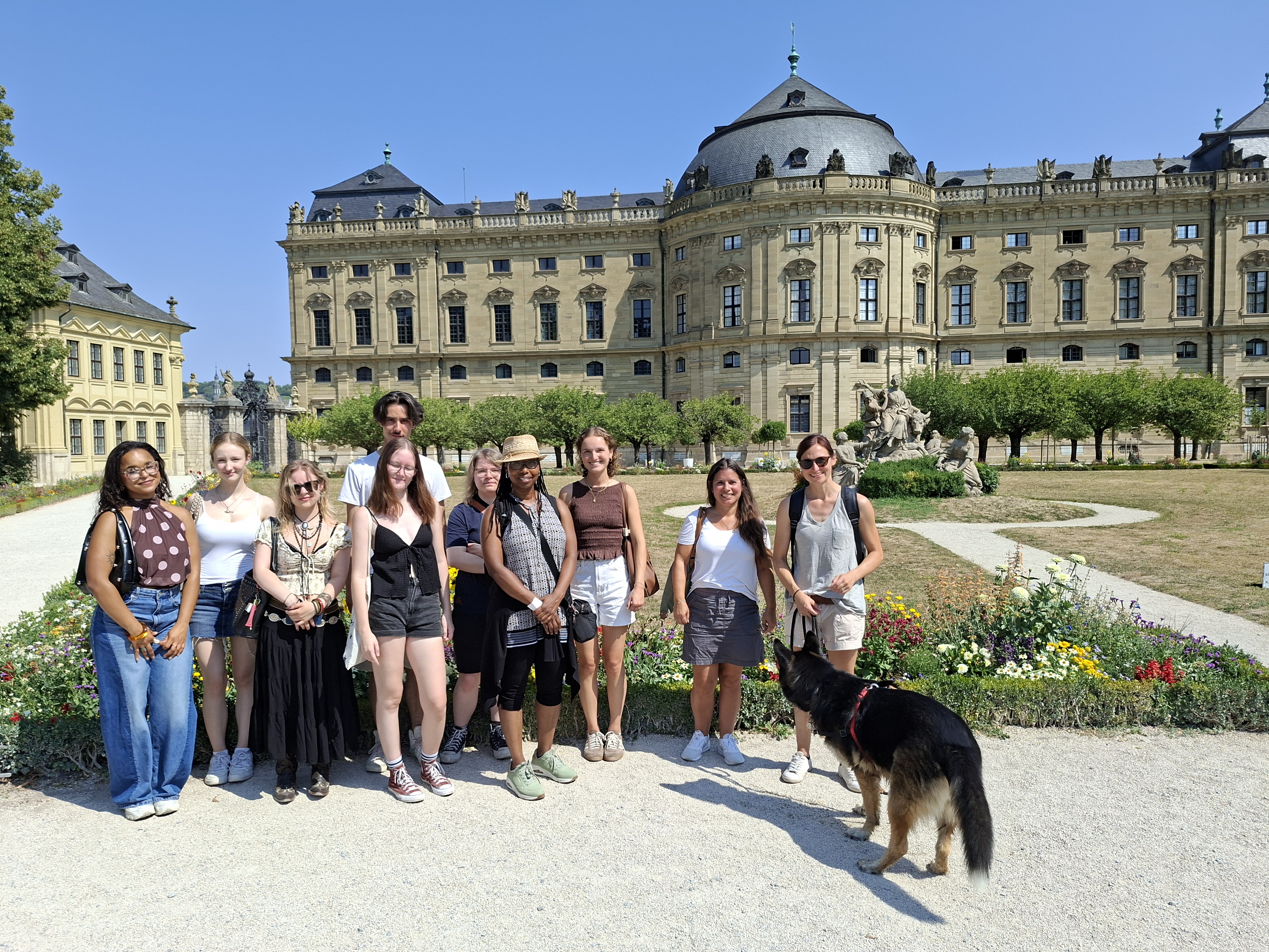 Eine Gruppe junger Erwachsener und Begleitpersonen posiert bei sonnigem Wetter in einer barocken Gartenanlage vor der Würzburger Residenz; im Vordergrund steht ein Hund.