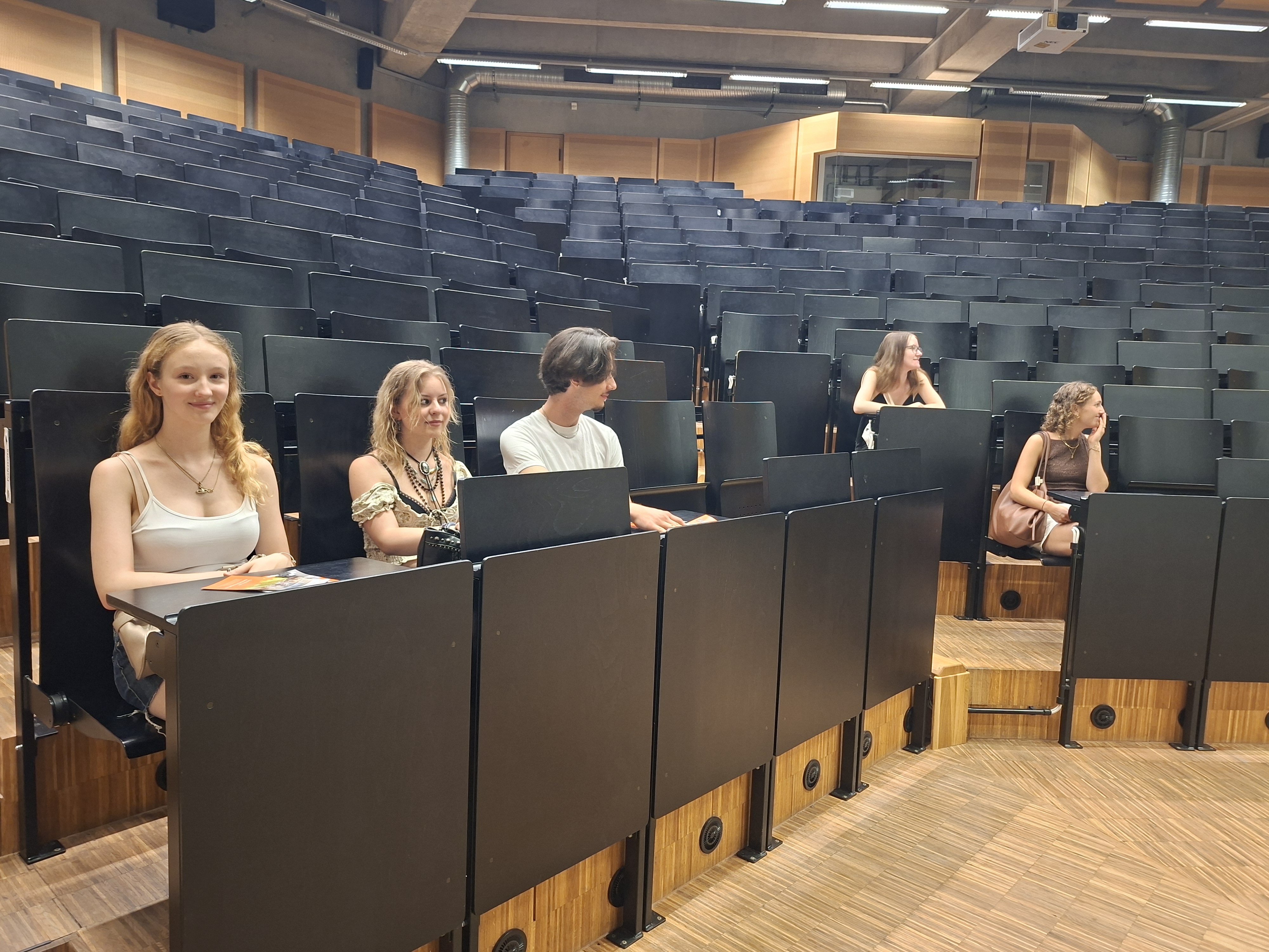 Mehrere junge Erwachsene sitzen verteilt in einem großen Hörsaal mit ansteigenden Sitzreihen.