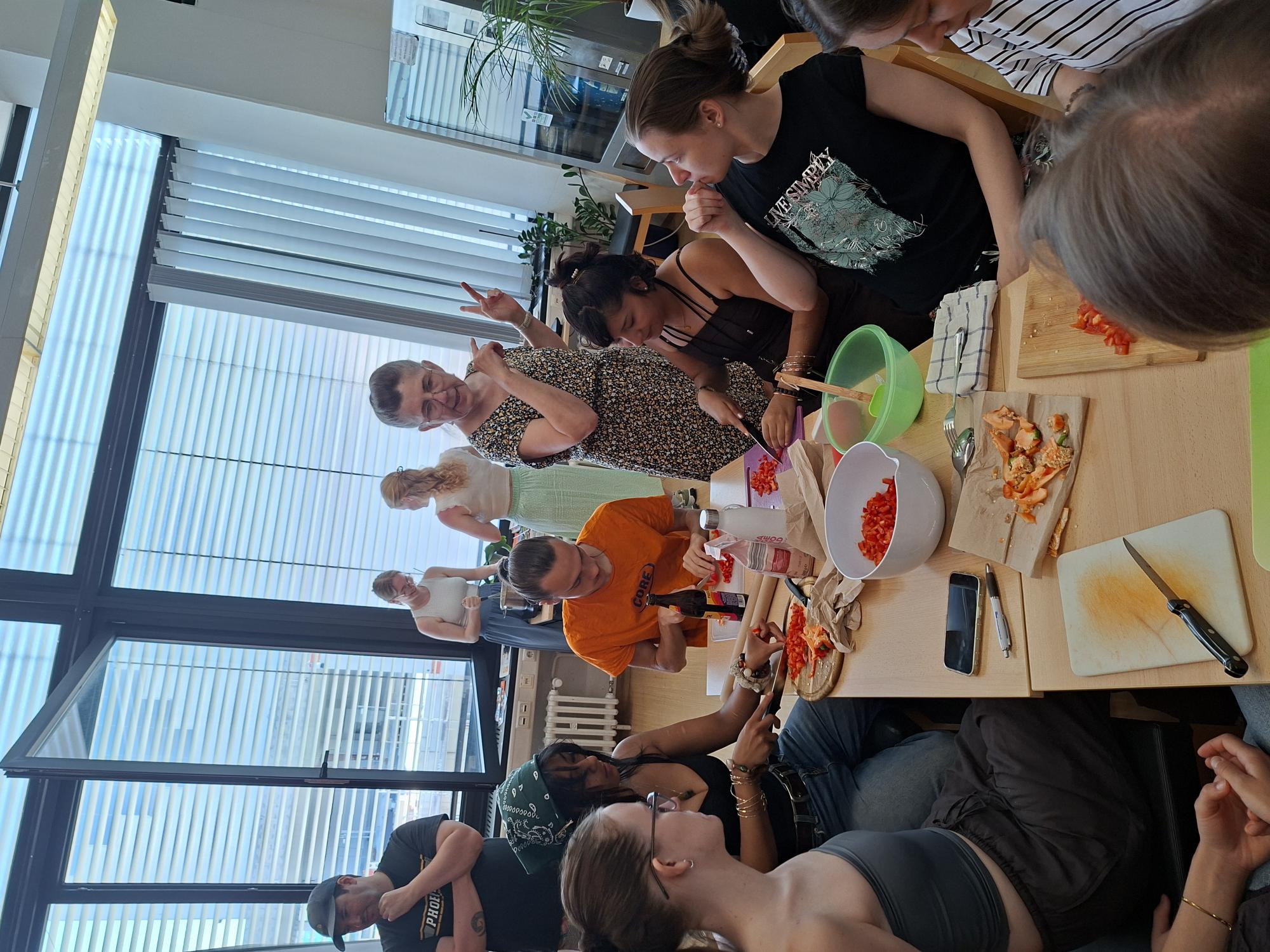 Eine Gruppe bereitet gemeinsam eine Mahlzeit zu und schneidet unter Anleitung einer Frau rote Paprika an einem großen Tisch.