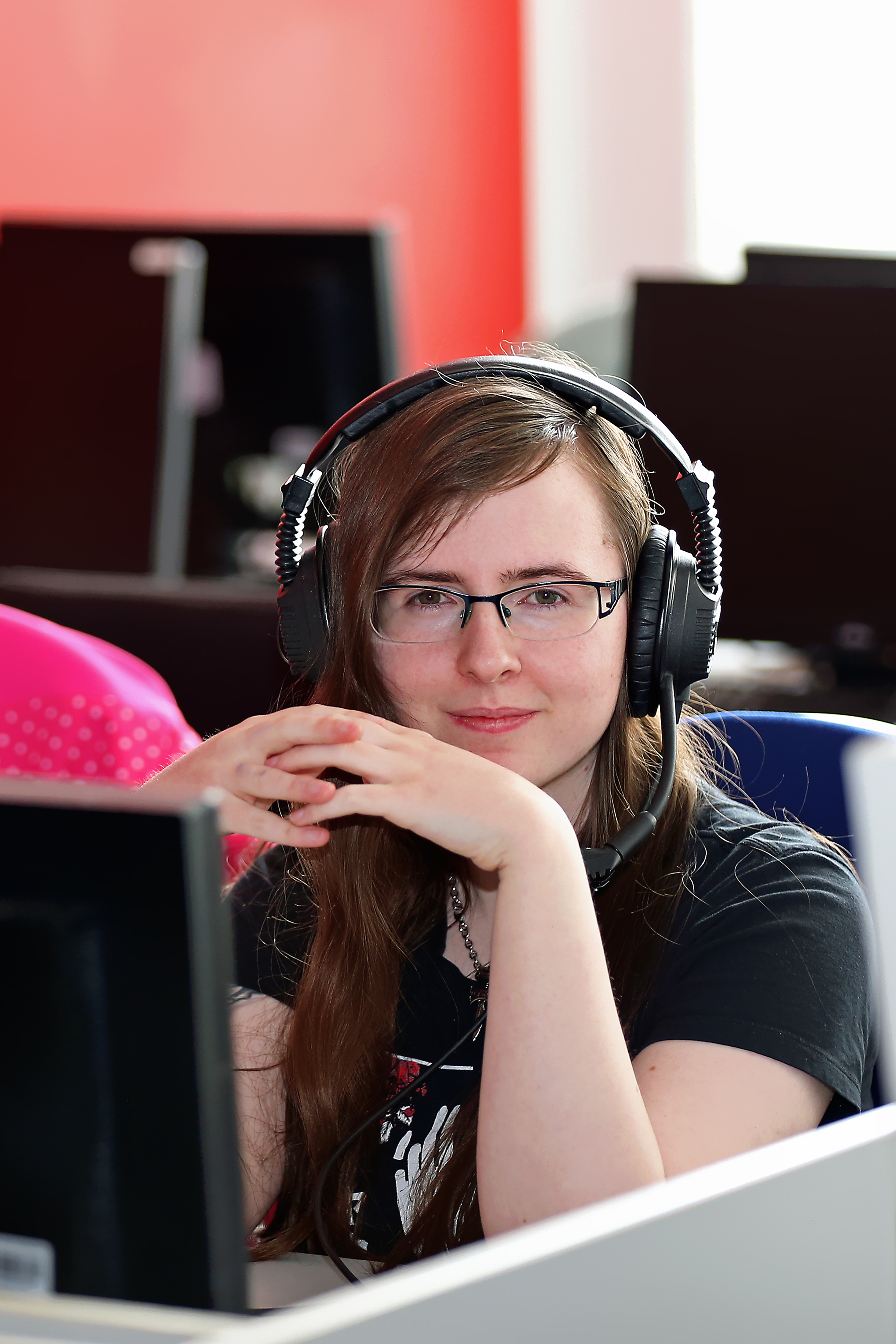 Schülerin mit langen Haaren, Brille und großem Headset sitzt an einem Computerarbeitsplatz, schaut direkt in die Kamera und verschränkt entspannt die Hände vor dem Gesicht.