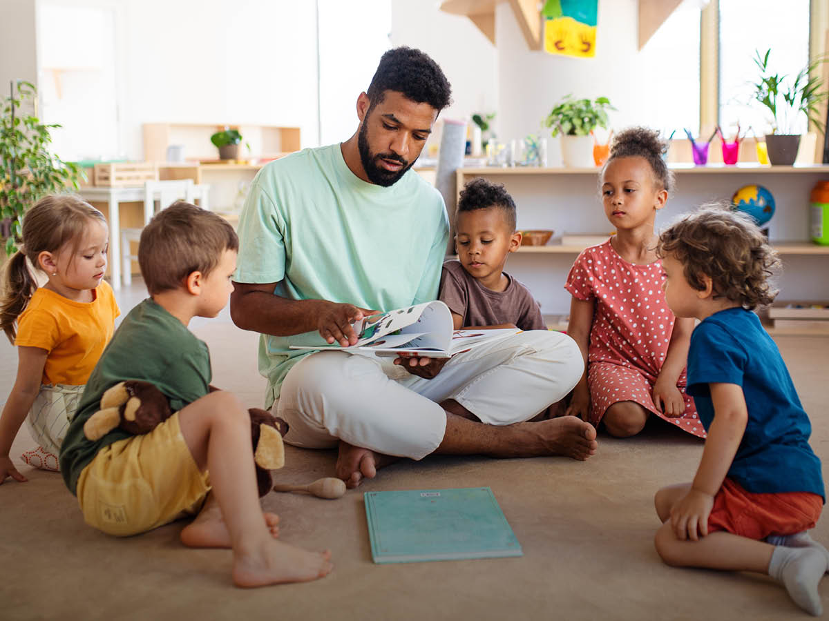 Eine erwachsene Person sitzt im Schneidersitz auf dem Boden und liest aus einem Buch vor, umgeben von fünf Kindern, die aufmerksam zuhören. Die Szene spielt sich in einem hellen Raum ab, der wie eine Kindertagesstätte aussieht.