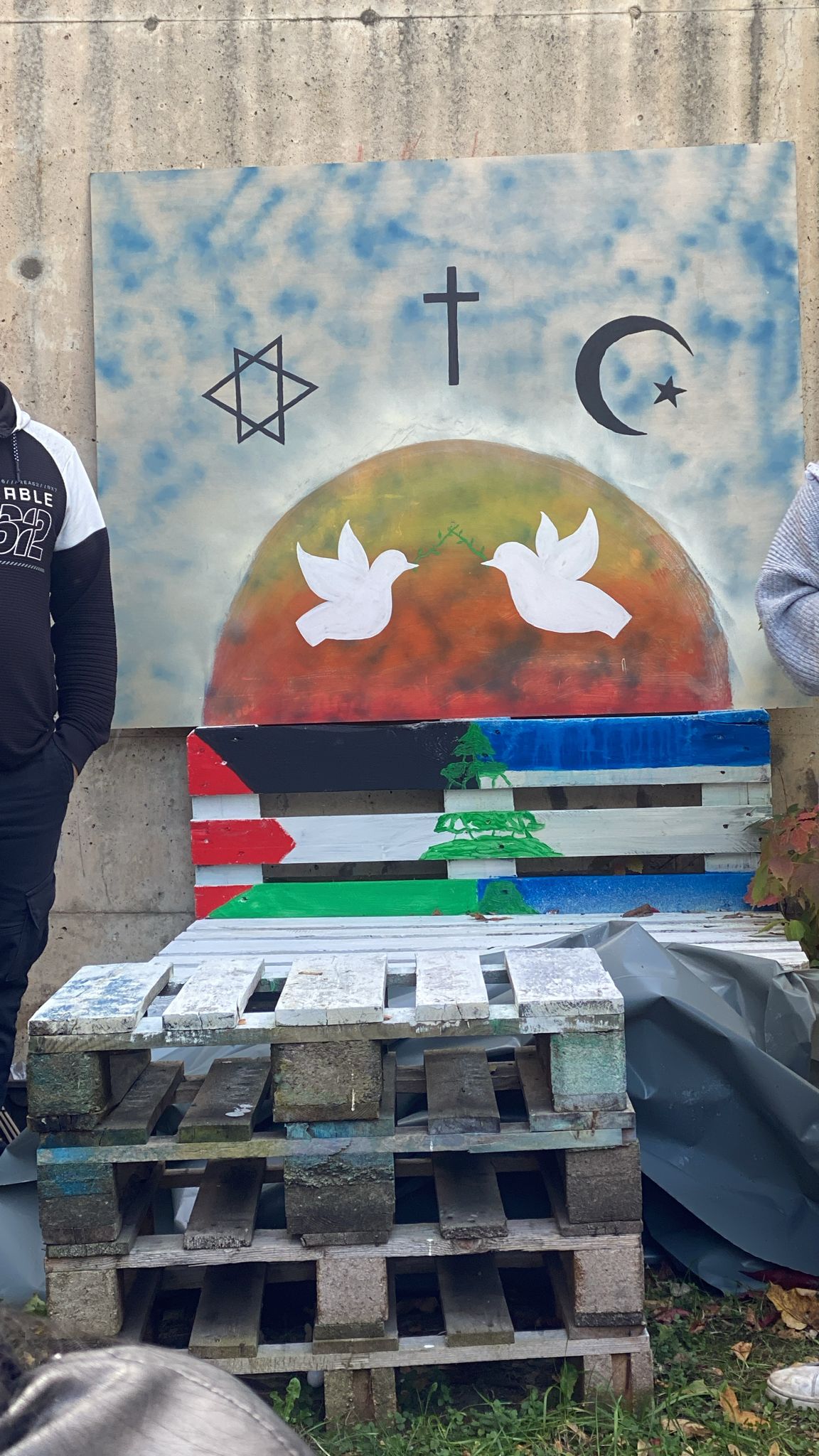 Partizipation und zeitgenössische Kunst Eine mit einer Israel-Flagge und Palästina-Flagge bemalte Holzpalette steht für einem Holzbrett, auf welchem eine Friedenstaupe und die Symbole des Christentums, des Islams und des Judentums aufgemalt sind.