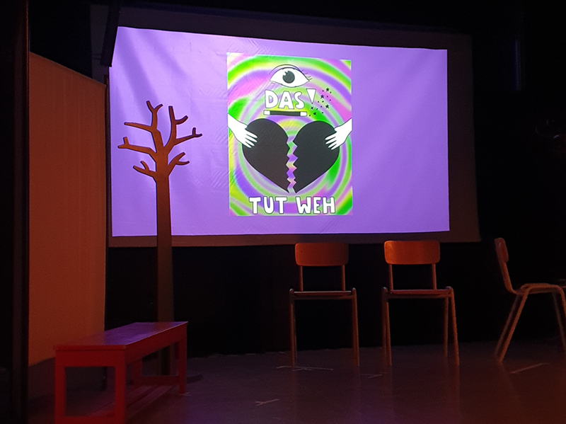 Das tut weh – Theaterprojekt setzt sich mit Erfahrungen auseinander Blick auf das Bühnenbild, mit roter Bank, drei Holzstühlen, einem Baum ohne Blätter, im Hintergrund der Titel Das tut weh an die Wand projiziert, mit Illustration eines gebrochenen Herzens.
